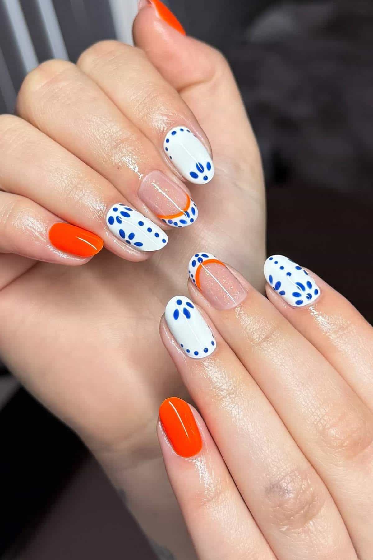 Hands with a manicure featuring solid orange nails, white nails with blue dotted patterns, and clear nails with orange and blue accents.