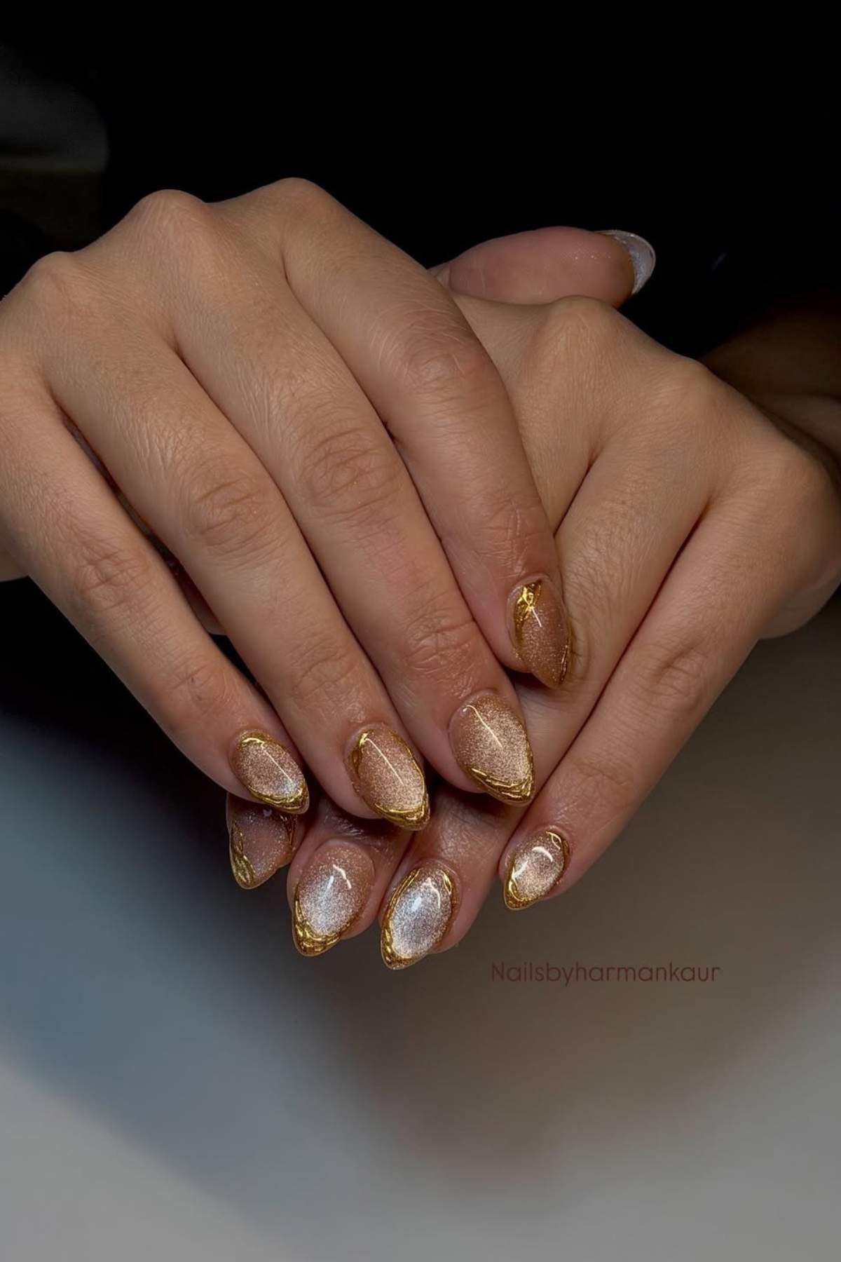 Hands with almond-shaped nails featuring a shimmery gold and silver design with gold accents.