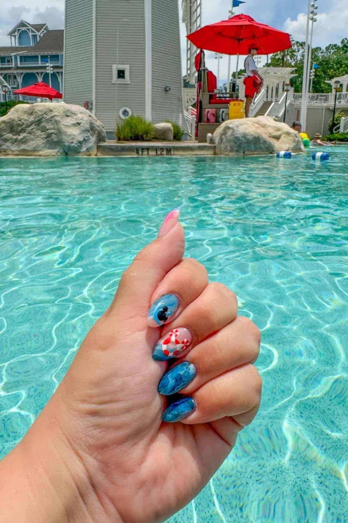 A hand with aquatic-themed nail art is held up over a swimming pool, with a lifeguard and poolside area visible in the background.