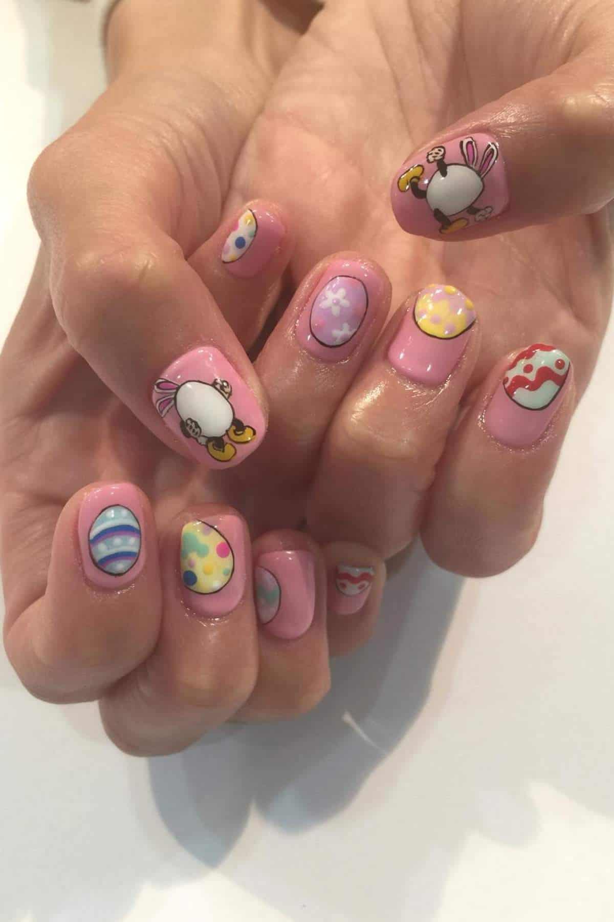Hands with short nails painted pink, featuring Easter-themed designs including eggs, polka dots, stripes, and cartoon bunny faces on each nail.