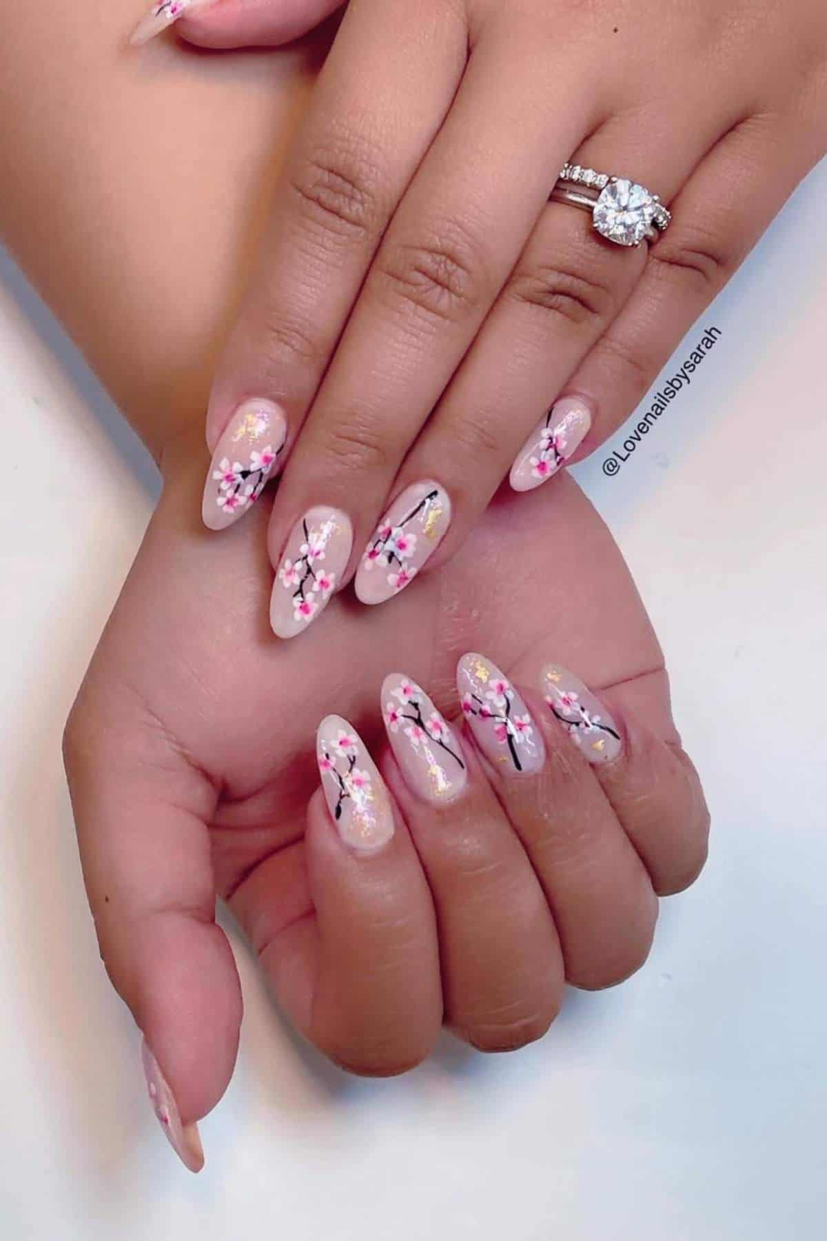 Hands with almond-shaped nails painted in a nude base and decorated with pink and white cherry blossom designs; wearer has a diamond ring on one finger.