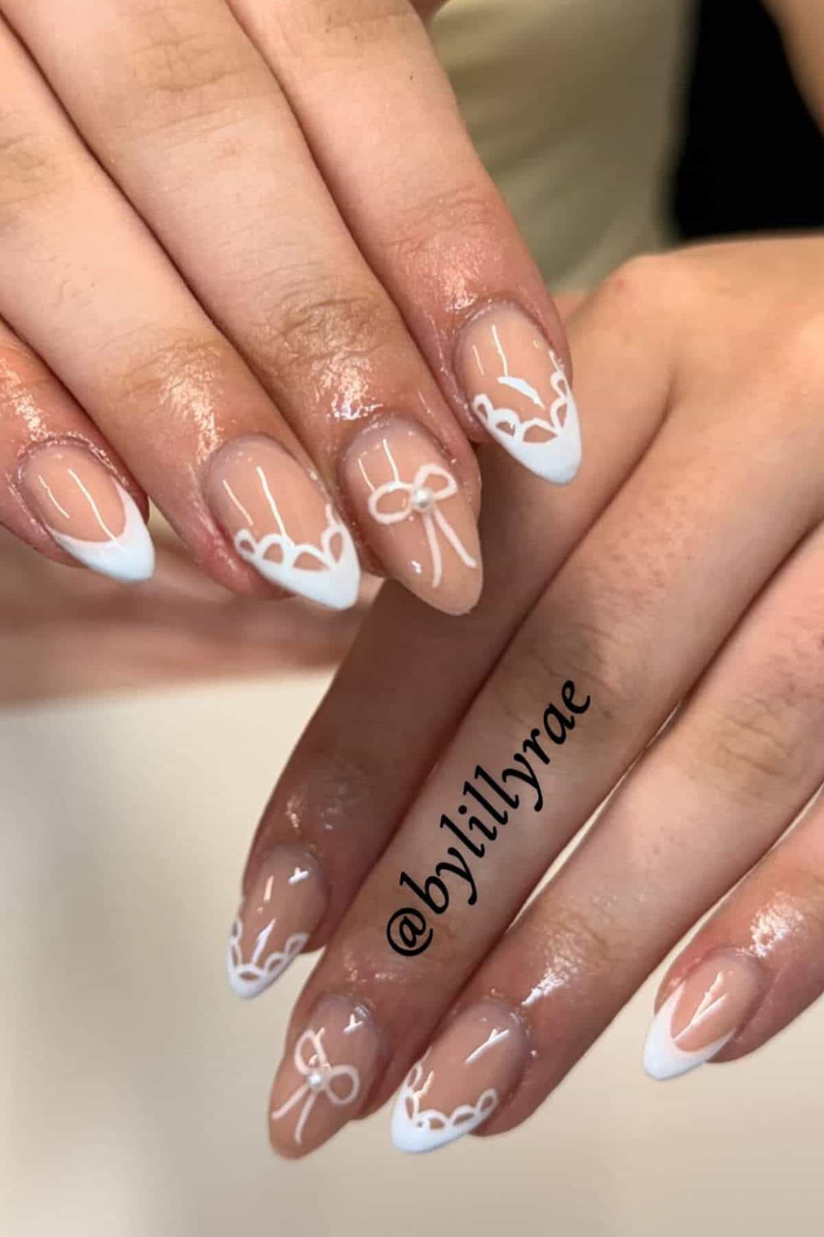 Close-up of hands with almond-shaped nails featuring a nude base, white French tips, and decorative white lace-like designs.