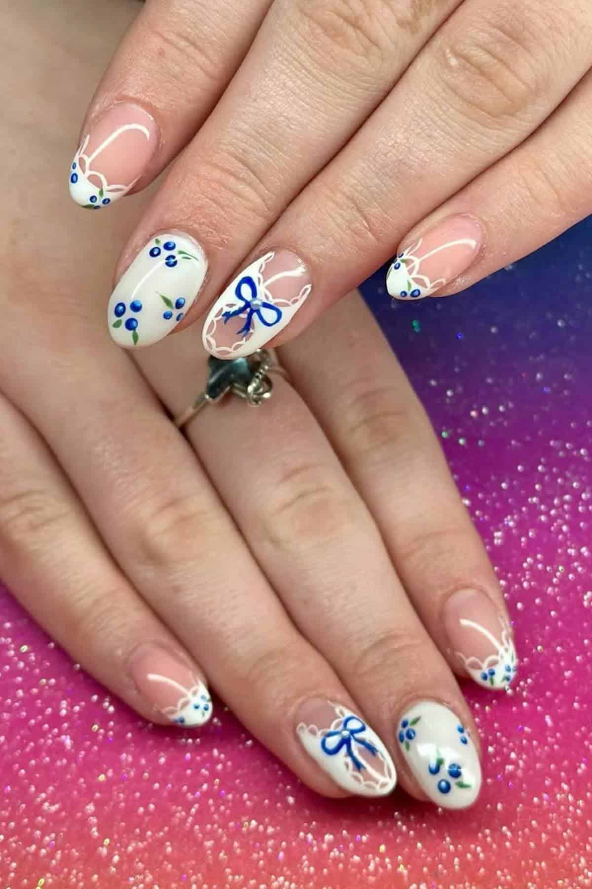 Hands with almond-shaped nails featuring white tips, blue flowers, and blue bow designs, displayed on a pink and purple gradient background.