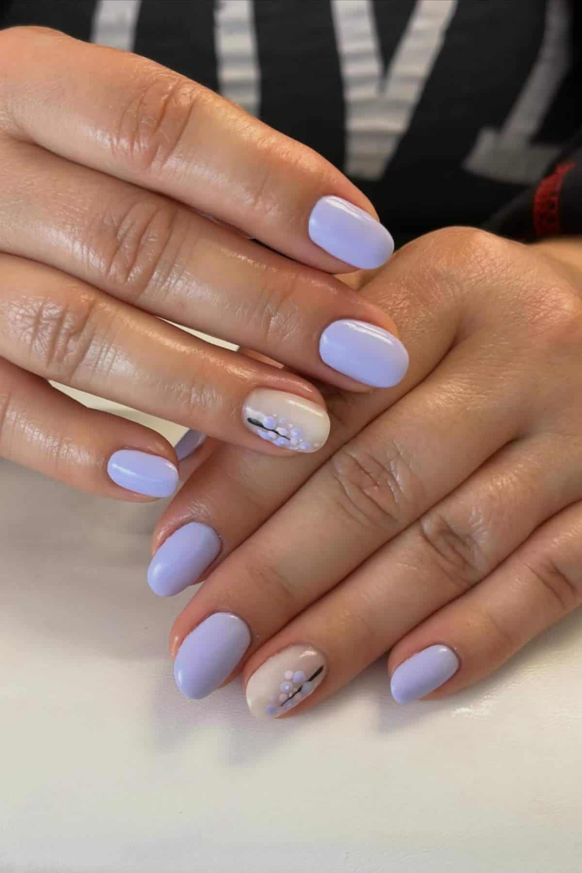 Hands with short, neatly manicured nails painted lavender, with accent nails featuring a nude base and small lavender and white floral design.