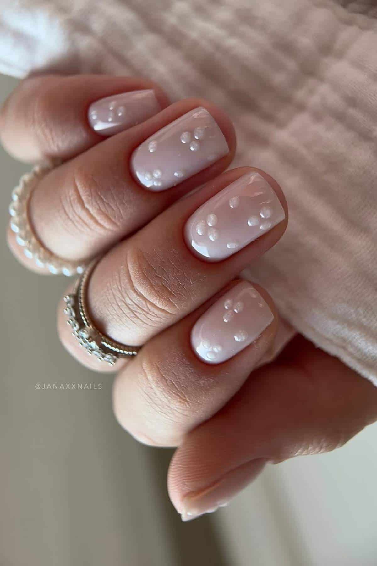 A hand with short, almond-shaped nails painted in pale pink polish with white 3D dot accents, wearing silver rings.