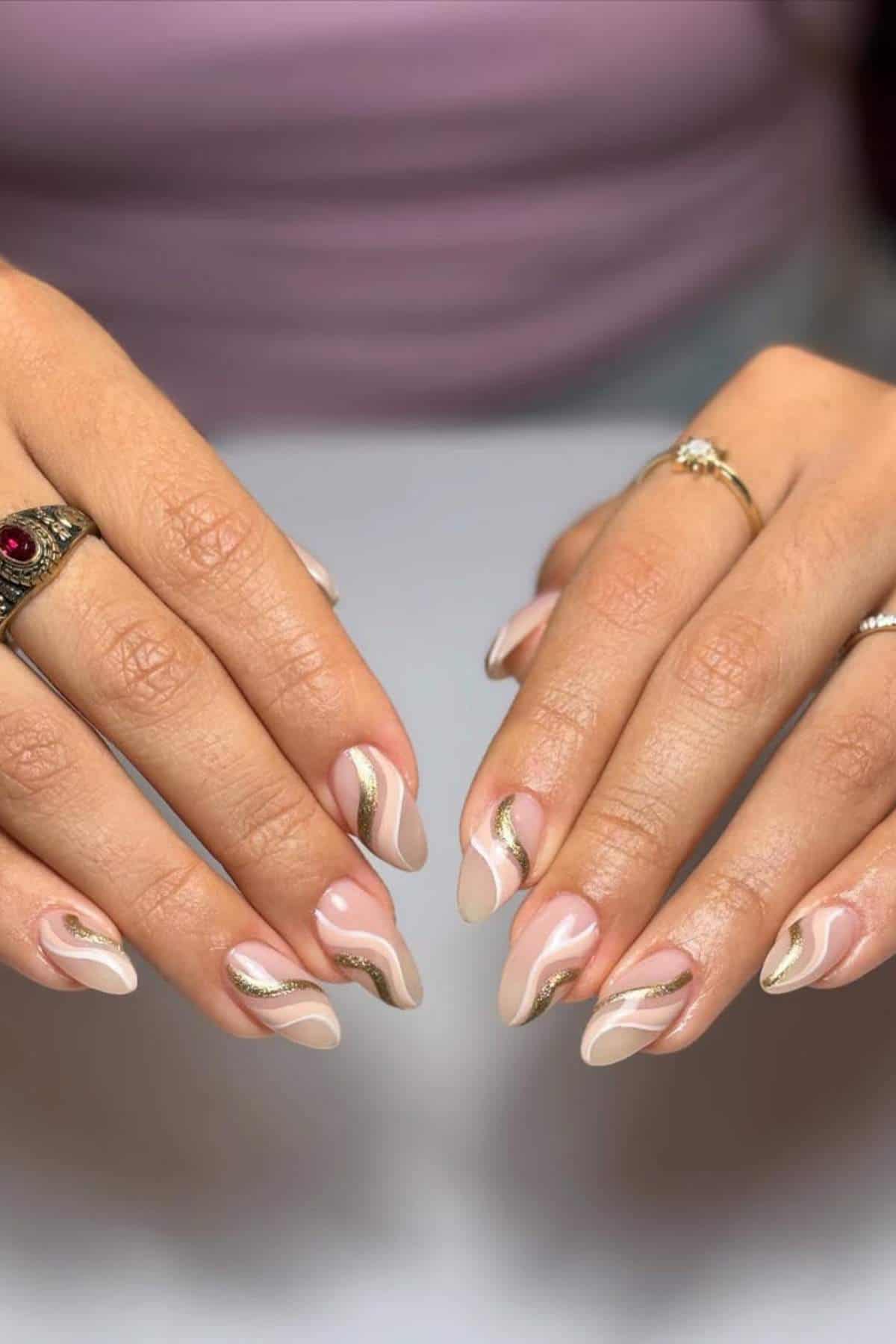 Hands with almond-shaped nails featuring a nude and white swirl design with gold accents. The person is wearing rings on both hands.