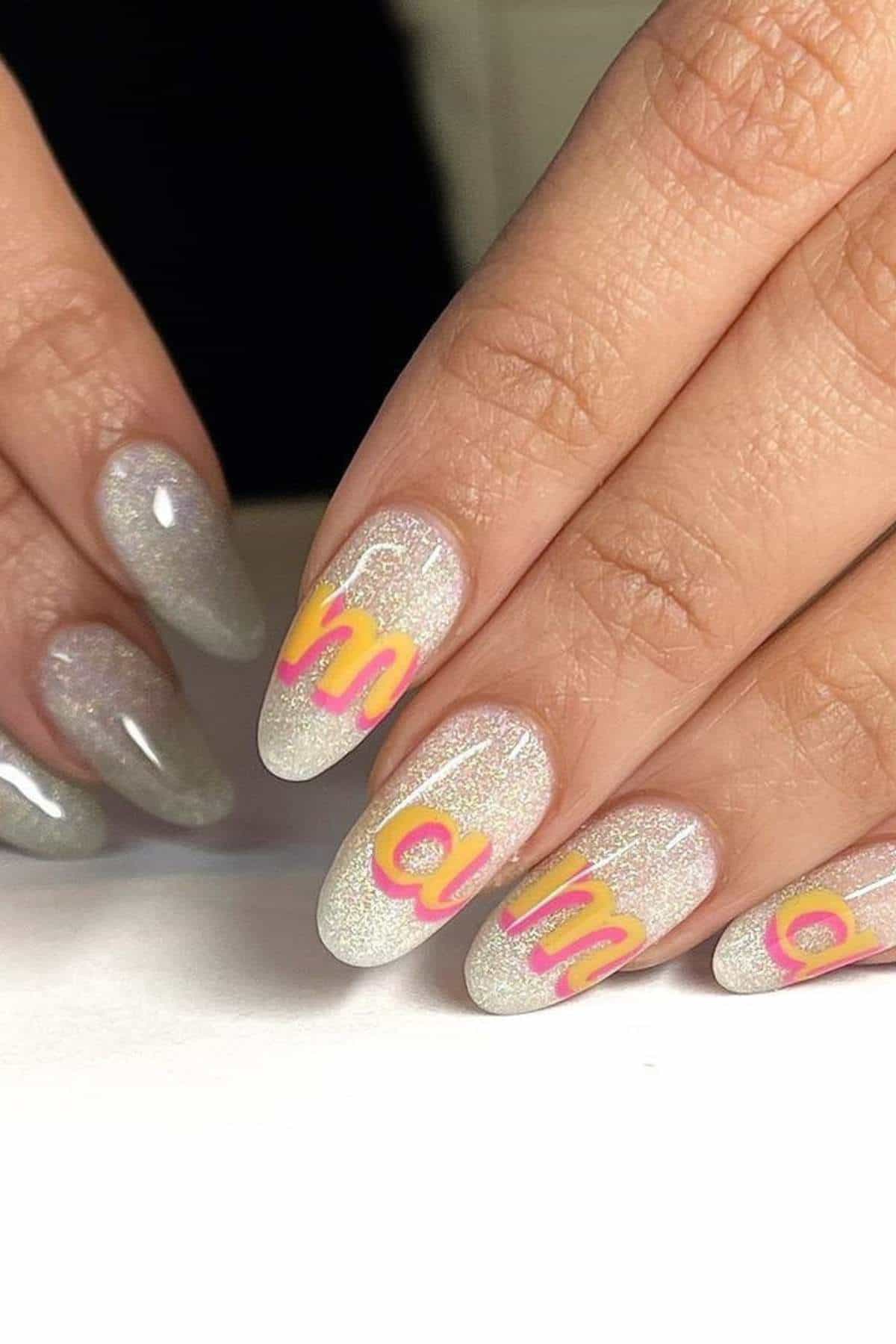 A hand with long, almond-shaped nails featuring a shimmery silver polish and bold letters in yellow with pink outlines spelling "mama" across four nails.