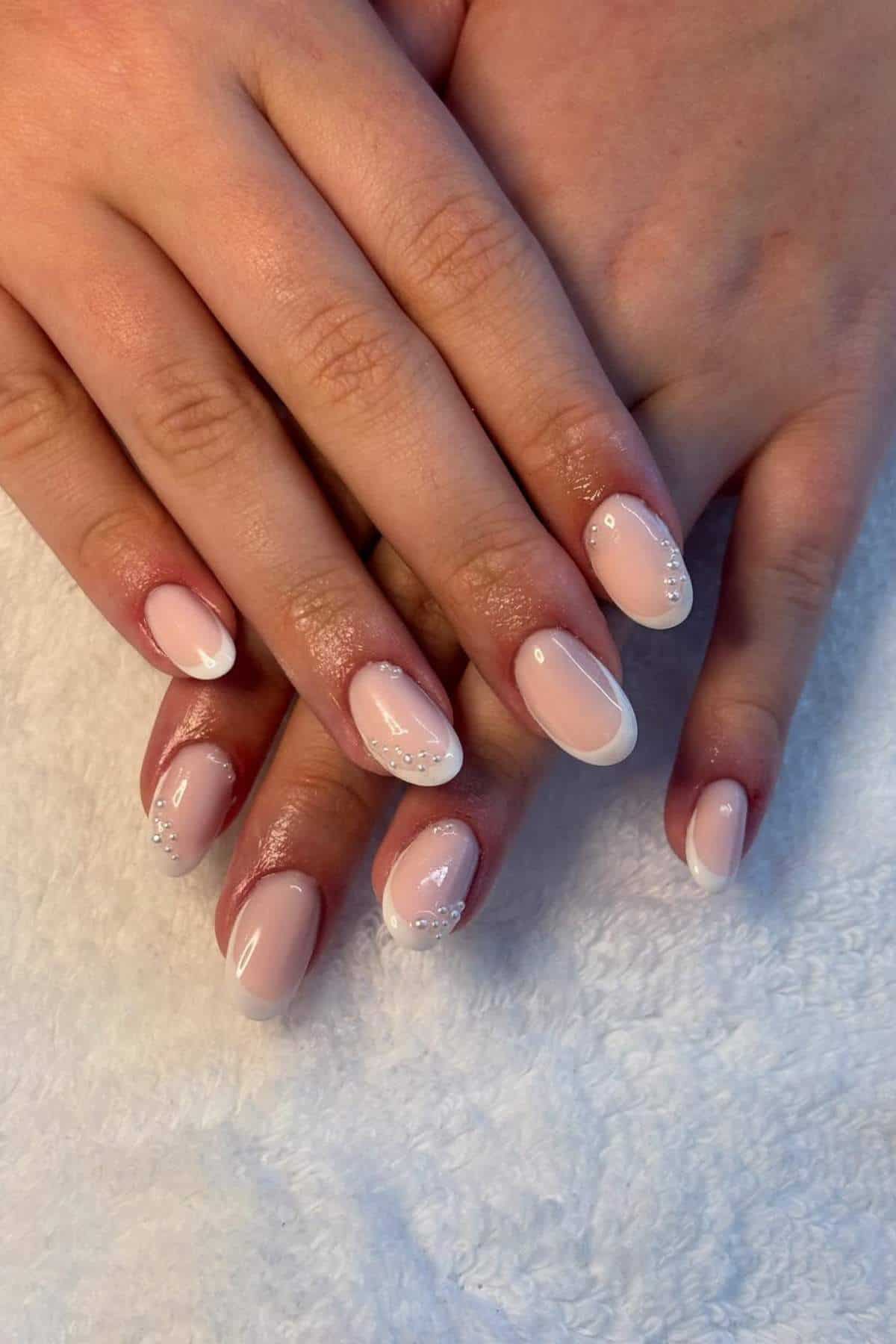 Hands with manicured nails featuring a pale pink base, white French tips, and small rhinestone accents, resting on a white towel.