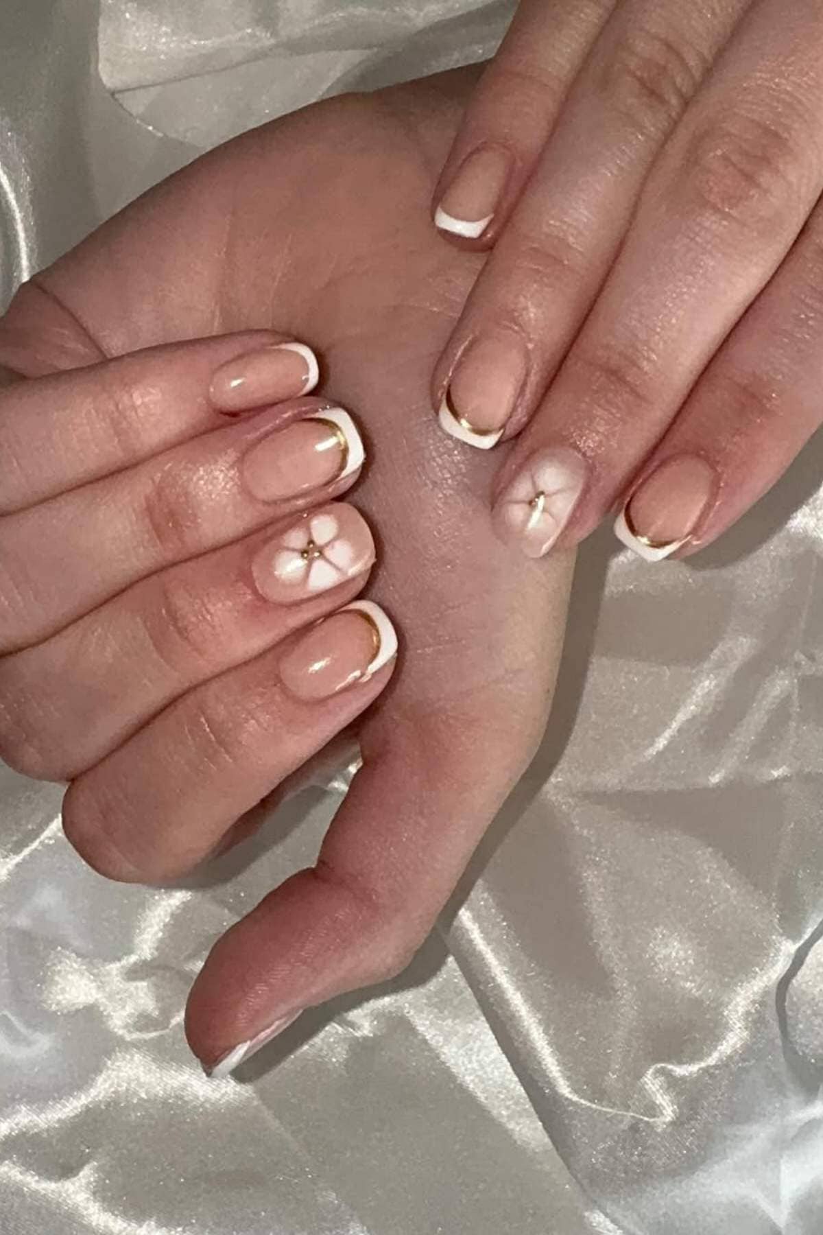 Hands with short, almond-shaped nails featuring a nude base, white French tips, gold accent lines, and a white flower design on two nails, shown against a satin background.