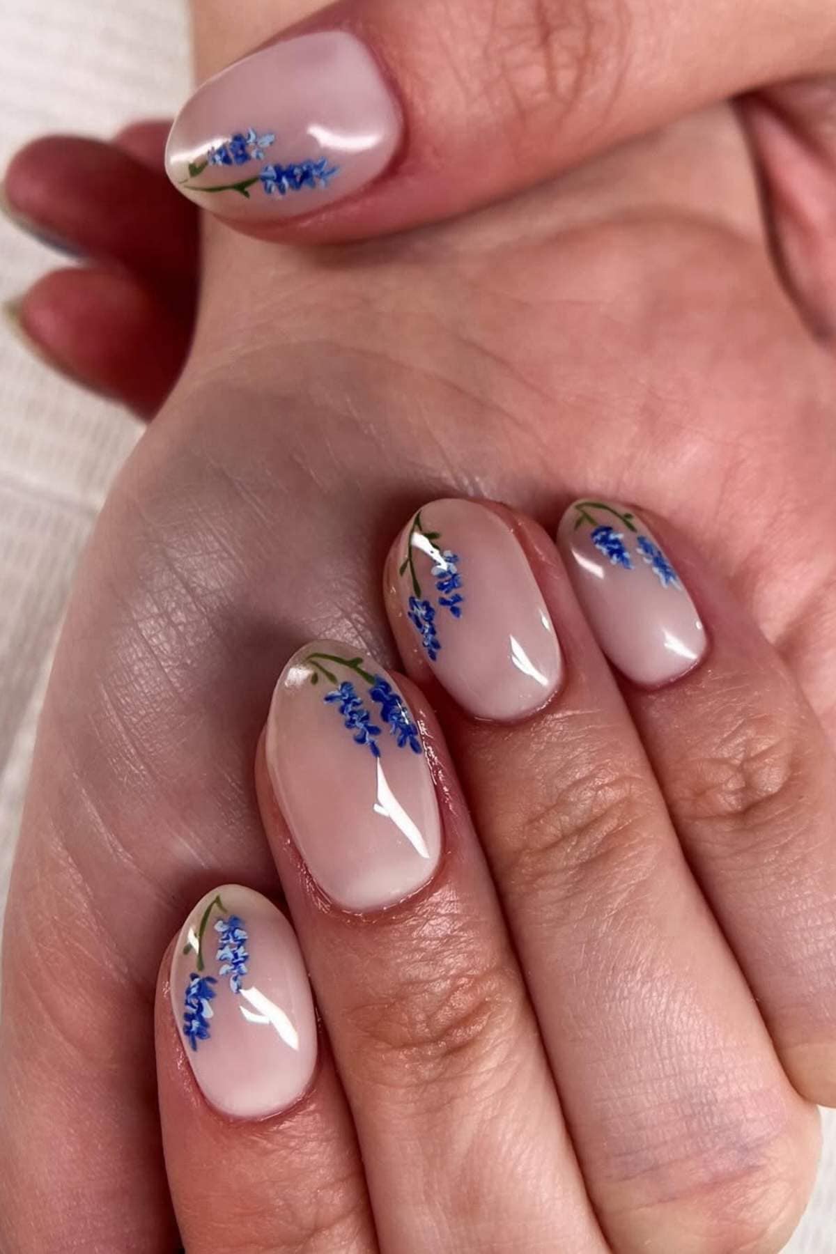 Close-up of hands with short, oval nails painted nude pink and decorated with small blue flower designs on each nail.