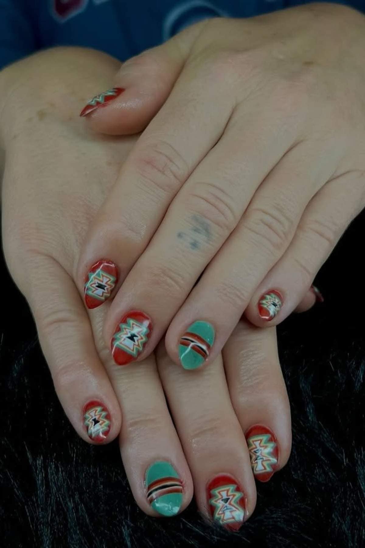 Hands with short, rounded nails featuring a geometric pattern in red, green, and white. The design has a Southwestern or Aztec-style motif on most nails.