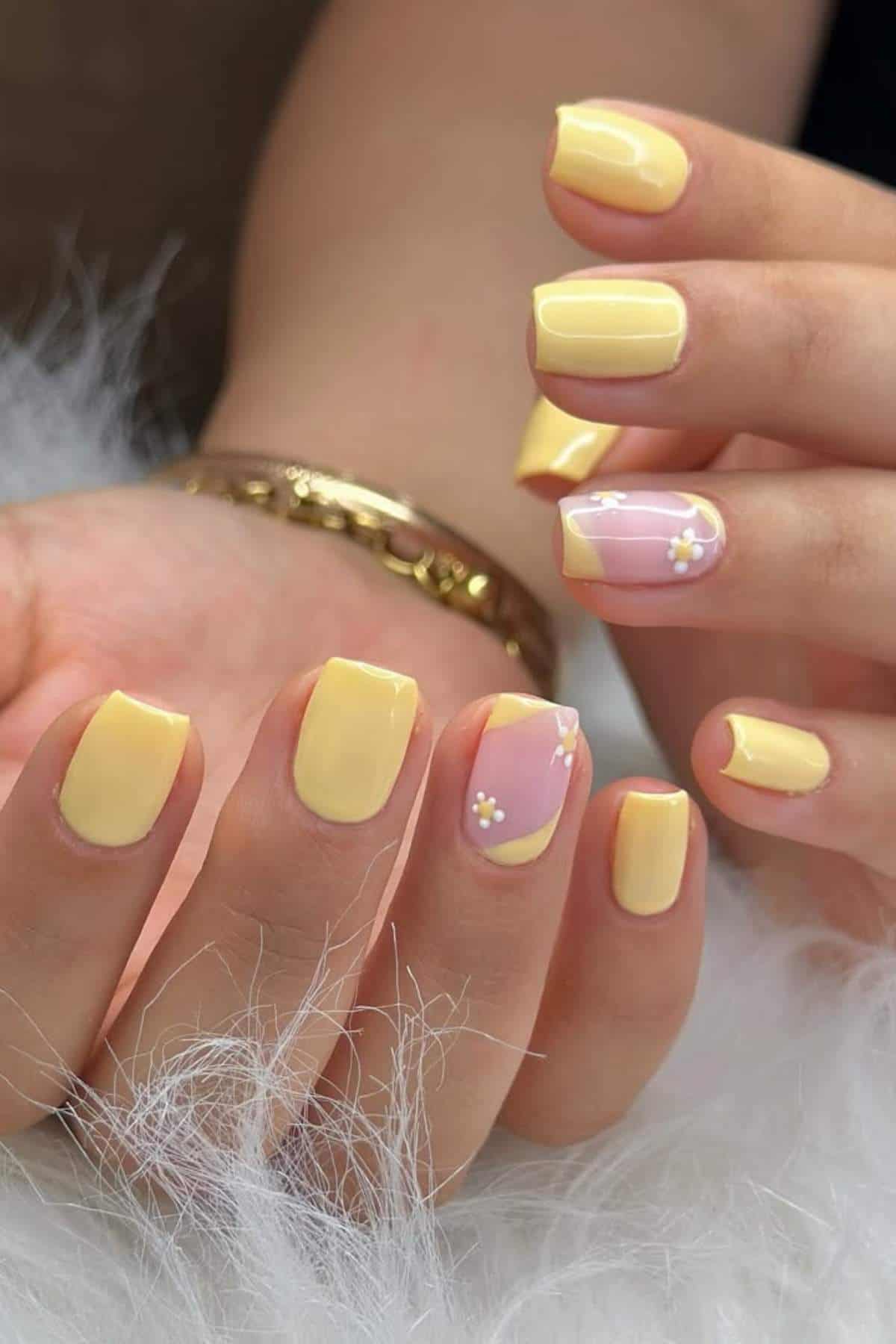 Close-up of hands with short, square-shaped nails painted yellow; two accent nails are pink with small white flower designs. A gold bracelet is on the wrist.