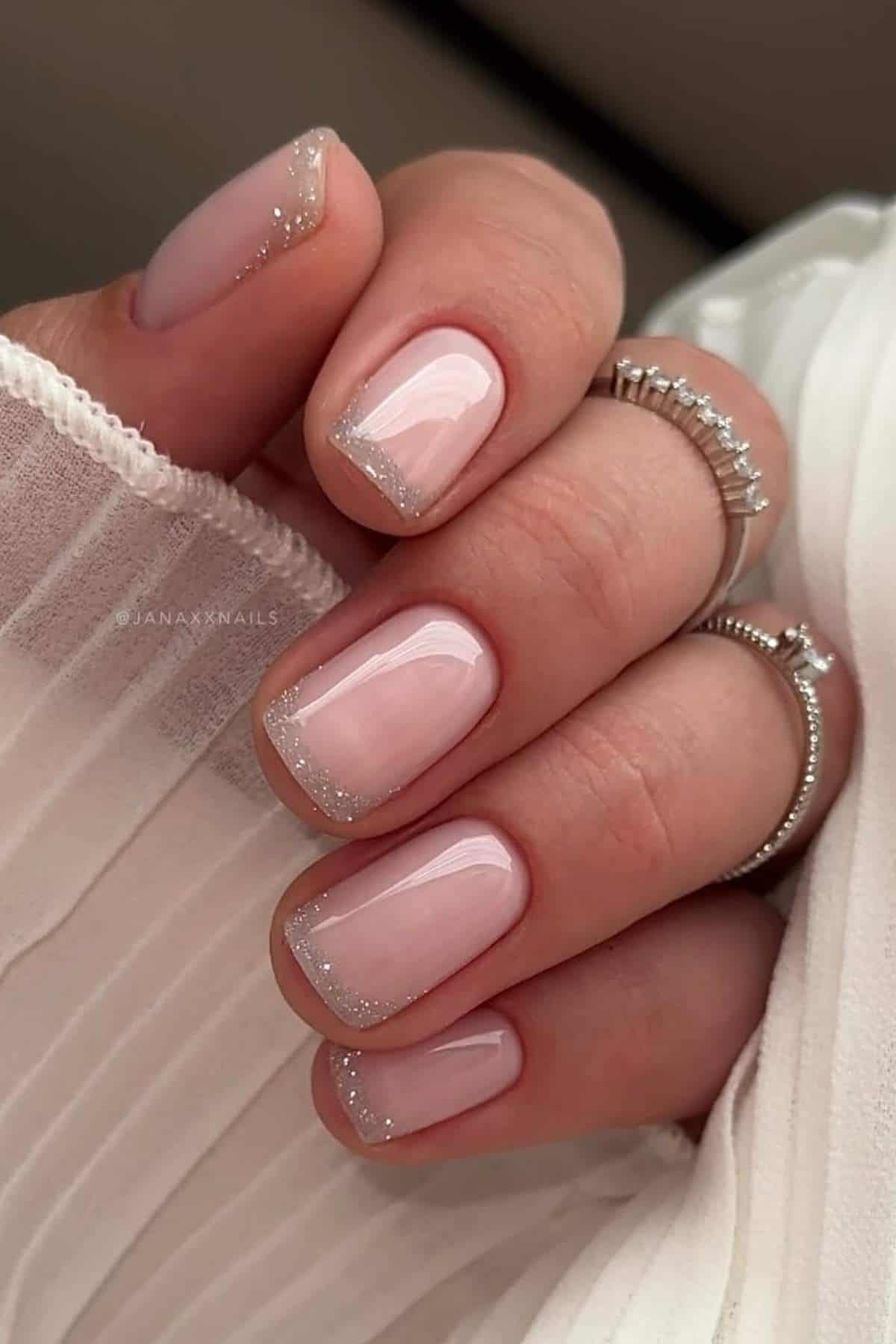 A hand with short, square-shaped nails painted in light pink polish with silver glitter tips, wearing silver rings and dressed in a light, textured fabric.