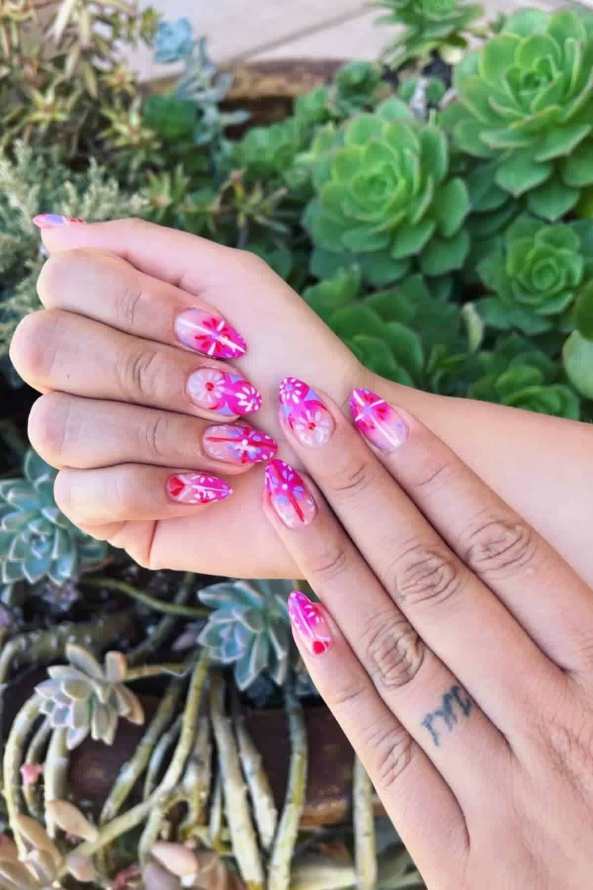 Hands with long, almond-shaped nails featuring pink and purple floral nail art, shown against a background of green succulent plants.