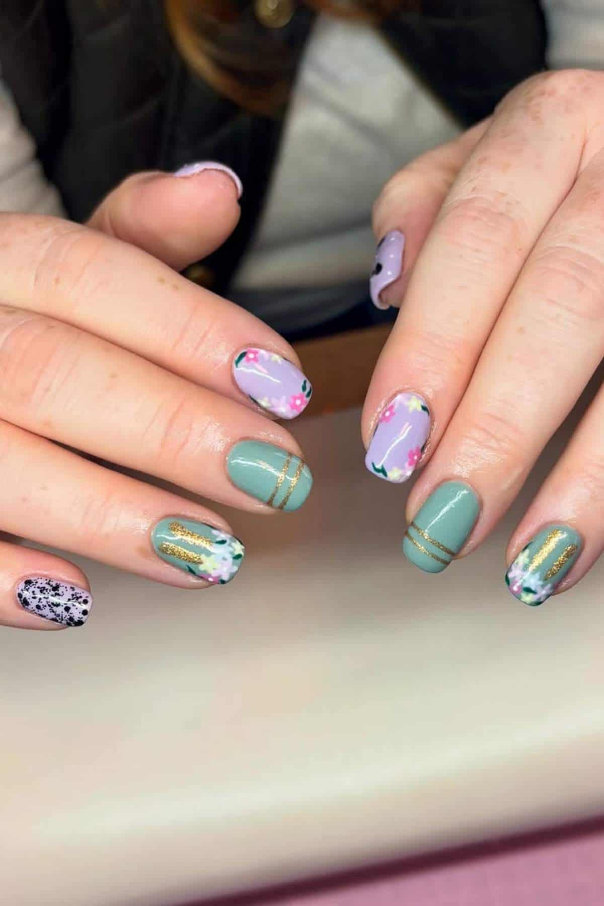 Hands with manicured nails featuring a mix of pastel colors, floral designs, gold stripes, and black speckled accents on each nail.
