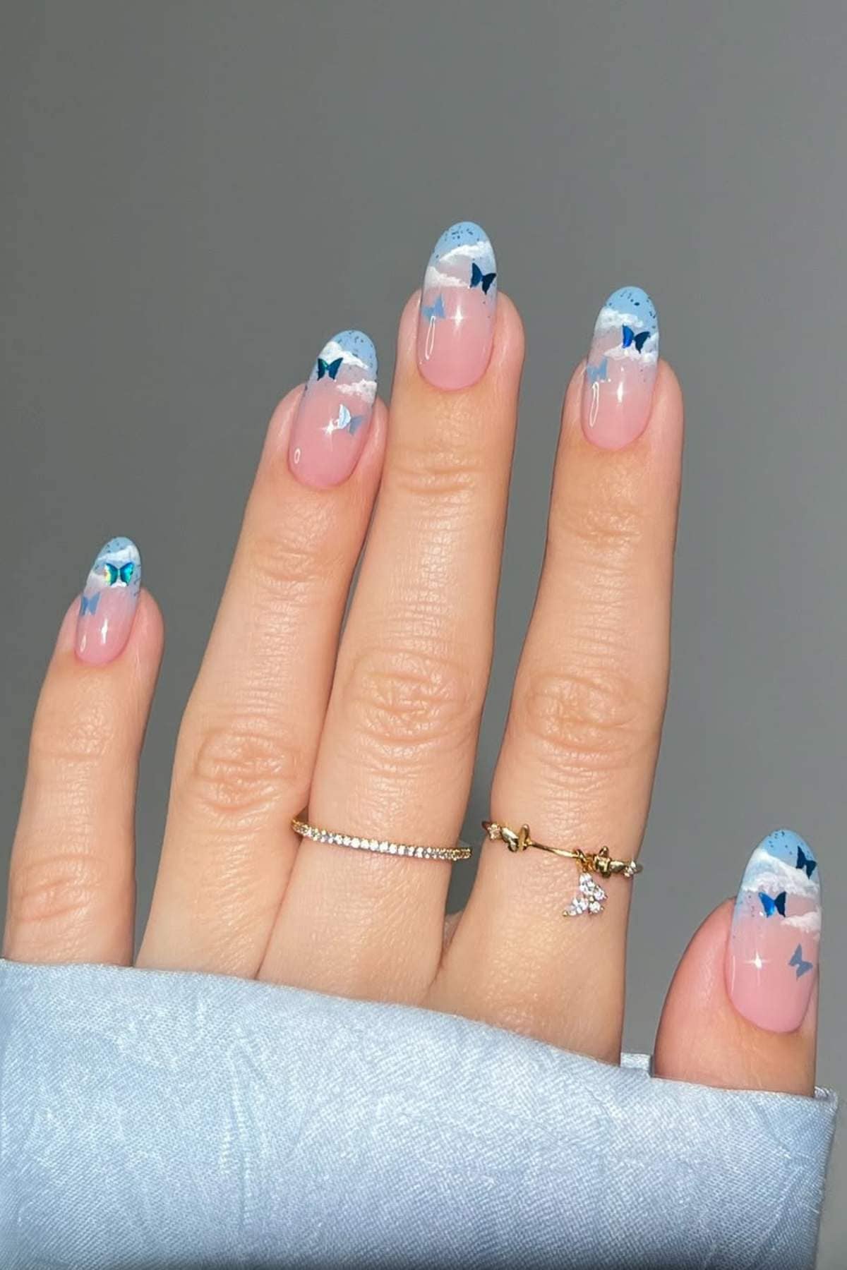A hand with long, almond-shaped nails featuring a sky-themed design with blue and white clouds and butterfly accents. The person is wearing two gold rings and a light blue sleeve.