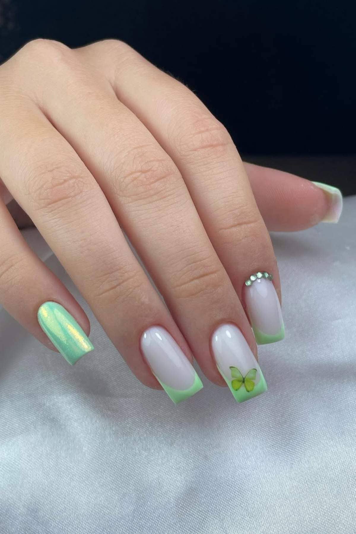 A hand with pastel green and white gradient manicure, featuring a butterfly design, small rhinestones, and a metallic green accent nail on a white surface.