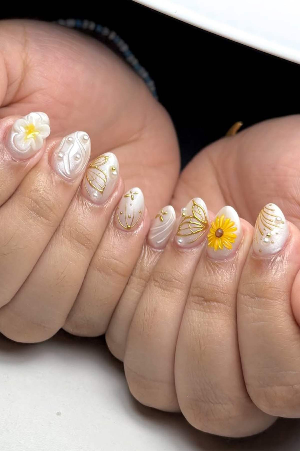 Close-up of hands with oval-shaped nails featuring white polish, yellow floral designs, gold accents, and 3D flower embellishments.