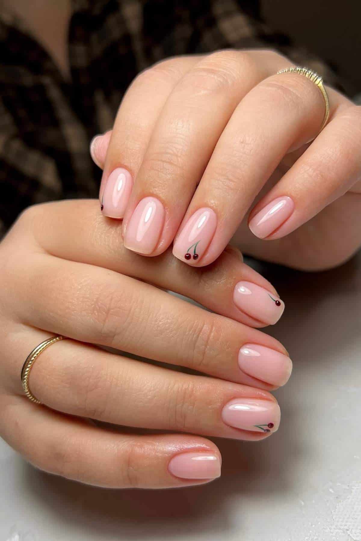 A close-up of hands with short, rounded nails painted light pink and small minimalist cherry designs on a few nails. The person wears thin gold rings on several fingers.