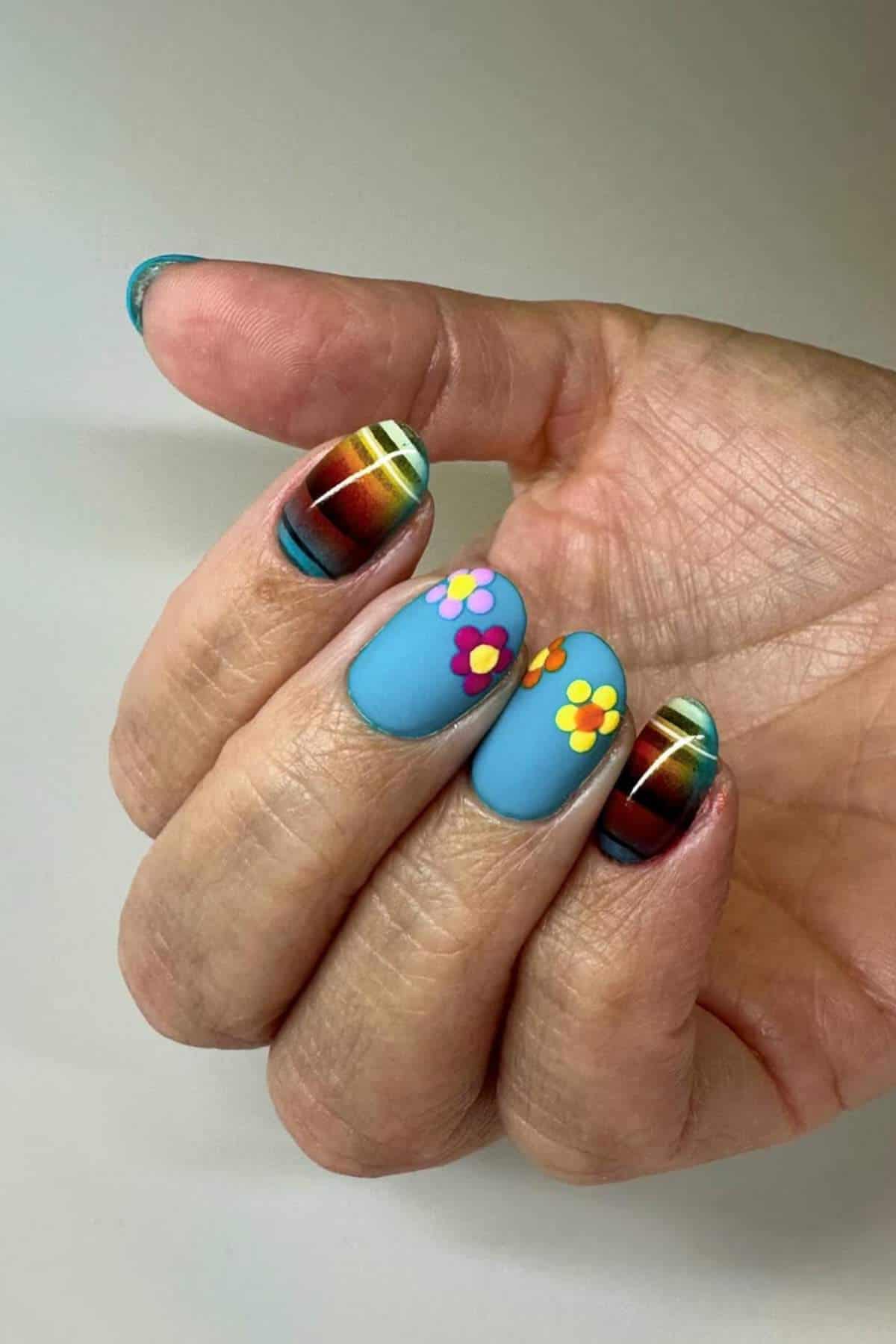 A hand with nails painted in blue with colorful flower designs on two fingers and multicolored striped patterns on the other nails.