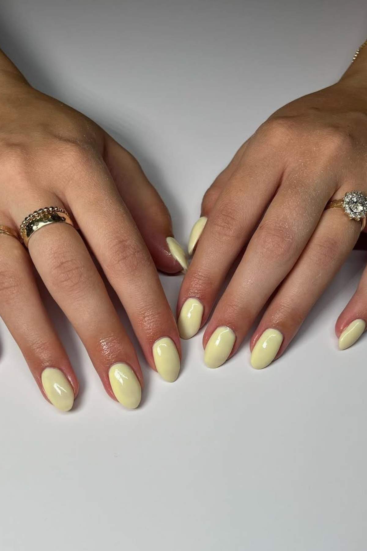 Two hands with oval-shaped nails painted in a pale yellow polish, resting on a white surface. The person is wearing two rings on their left hand and one on their right hand.