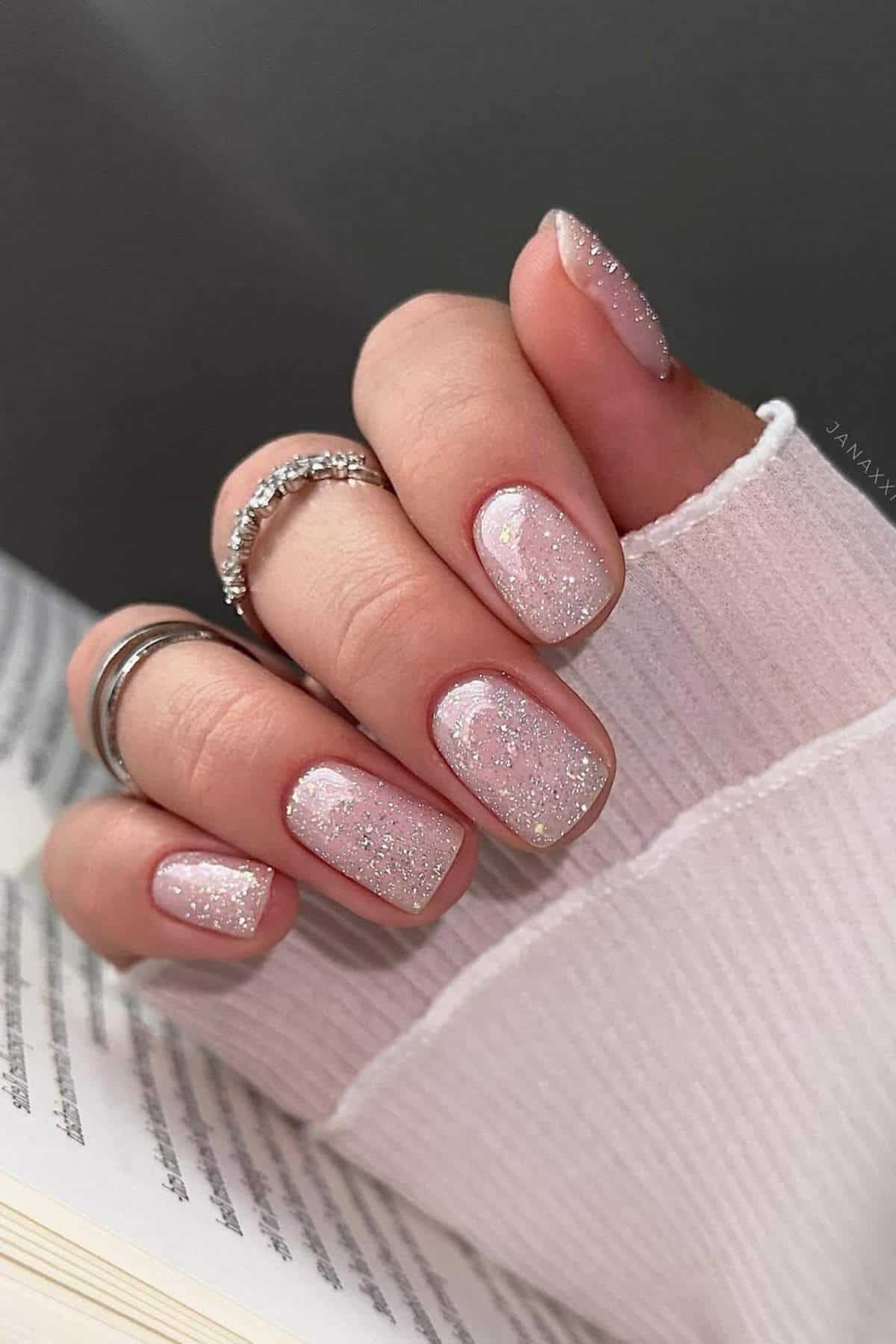 A hand with short, square-shaped nails painted with light pink glitter polish, wearing silver rings, rests on an open book.