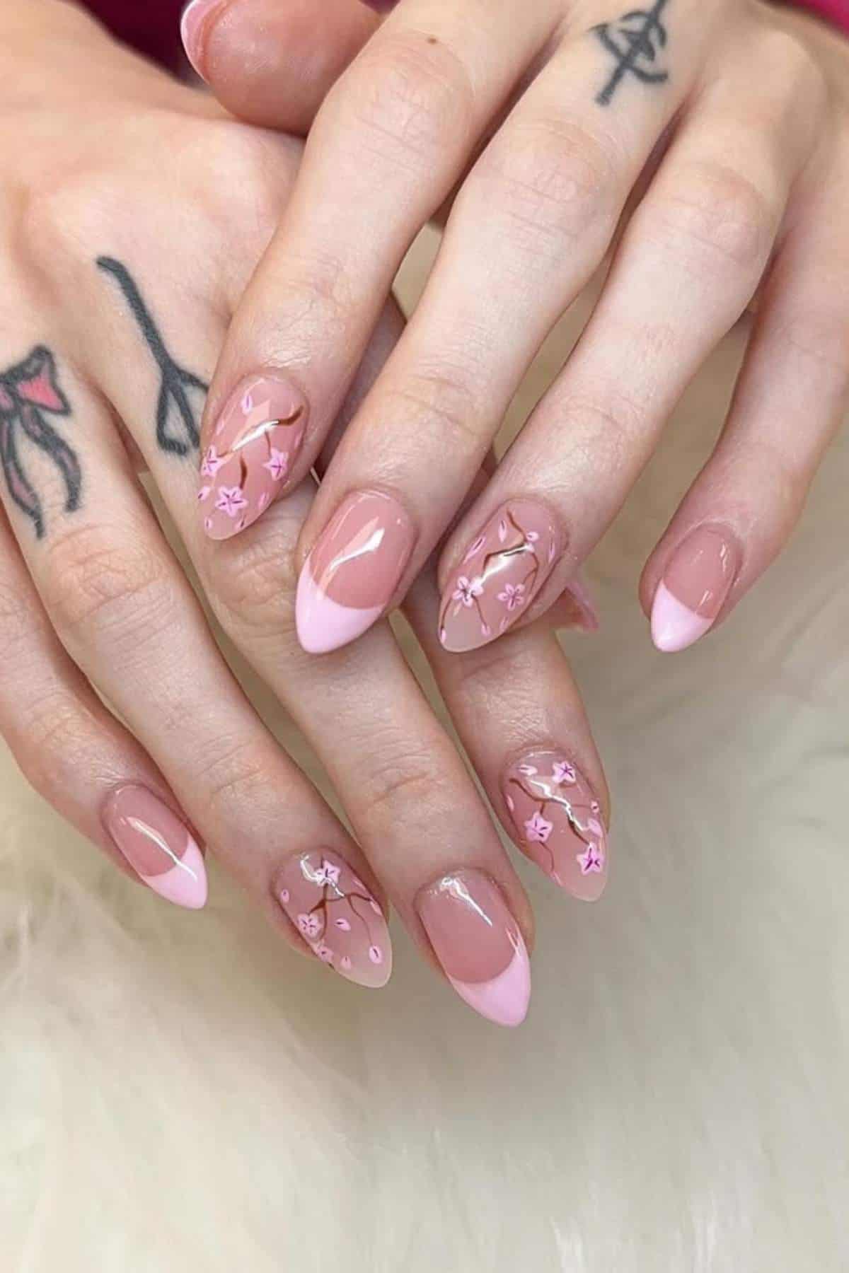 Hands with tattoos showing almond-shaped nails featuring pink French tips and floral designs on a light nude base.