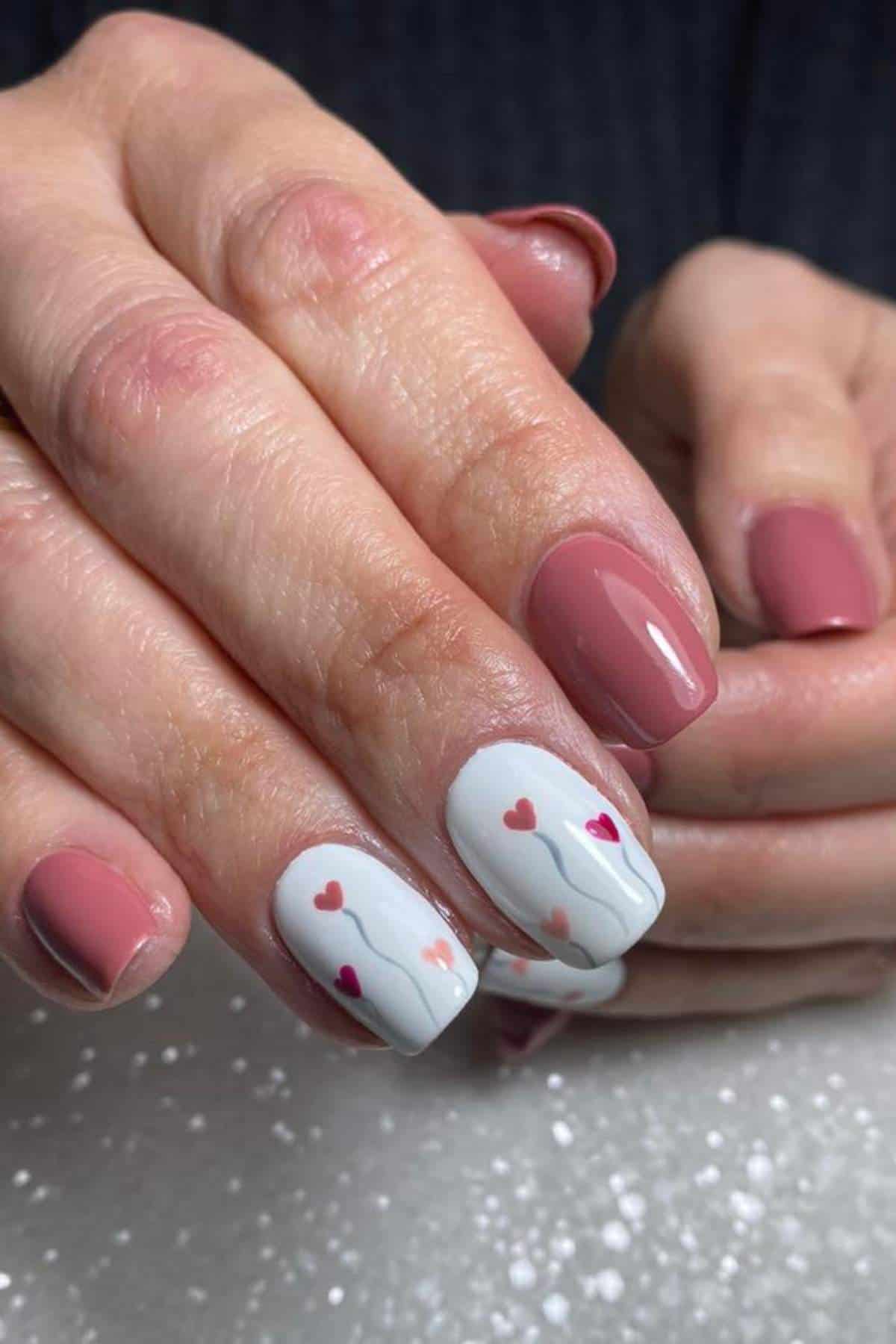 A hand with short, rounded nails painted pink, with two accent nails featuring a white base, small red and pink hearts, and thin wavy lines.