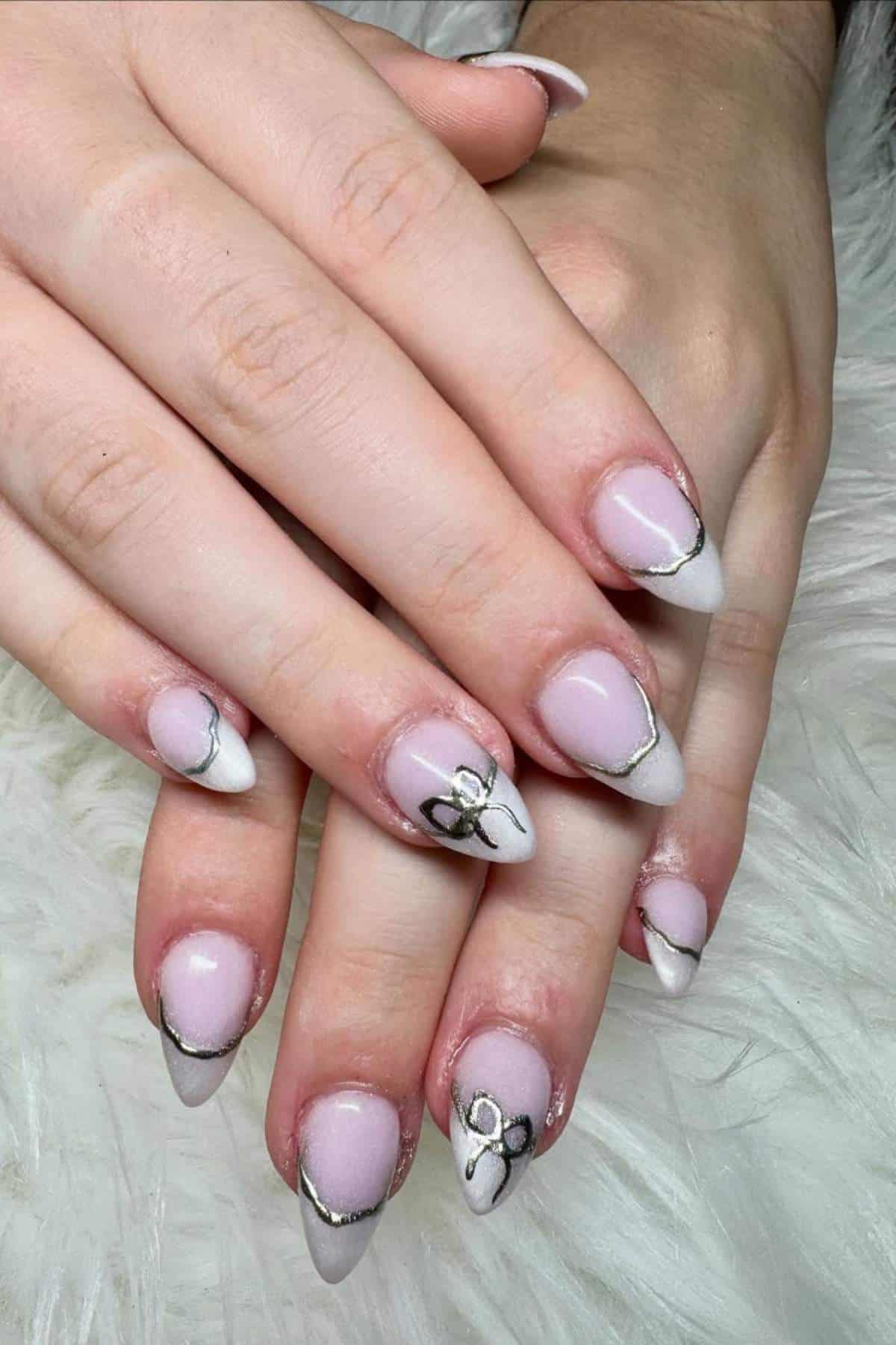 Hands with almond-shaped nails featuring a pale pink base, white tips, gold outline accents, and black and gold floral designs on some nails, resting on a white textured surface.