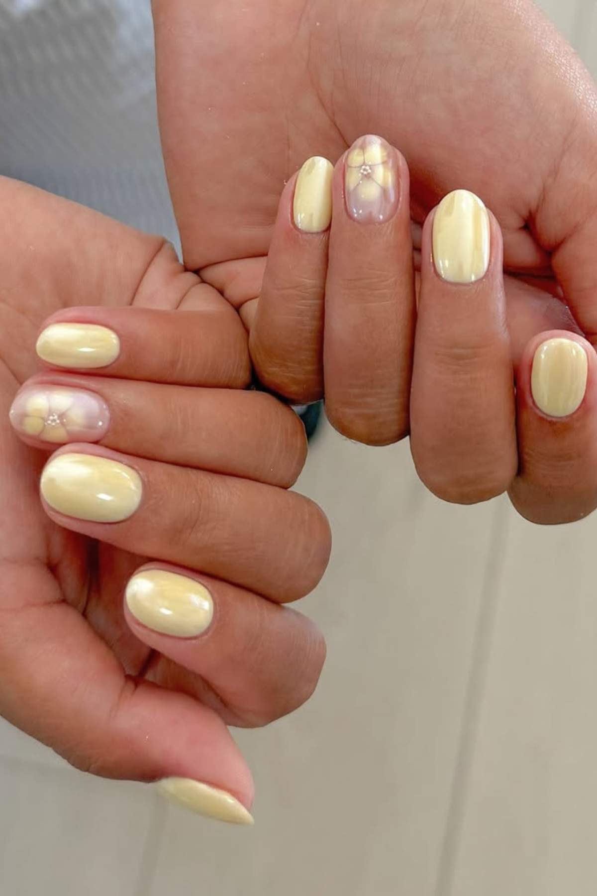 Hands with short, oval-shaped nails painted pale yellow; some nails feature a translucent base with subtle yellow flower designs.