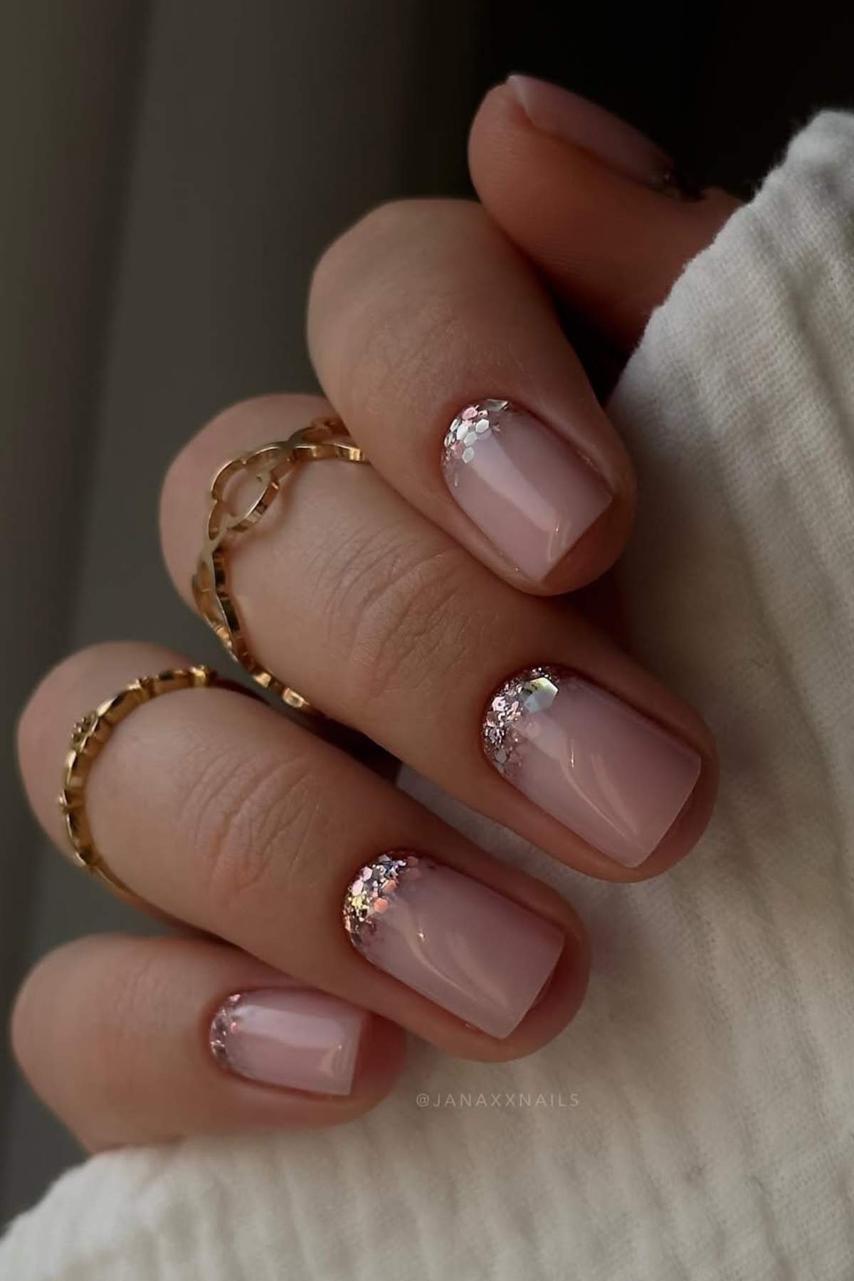 A hand with short, square-shaped nails painted in light pink polish with silver glitter accents near the cuticles; the person wears gold rings.