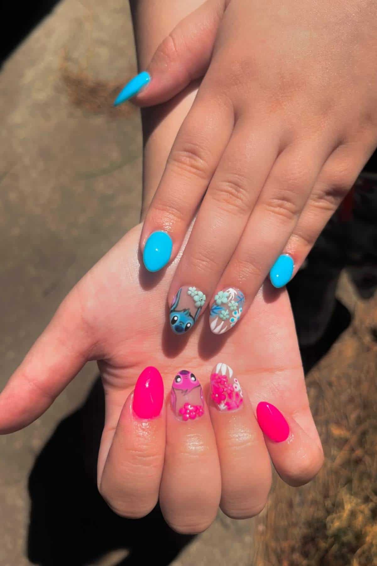 Two hands with manicured nails; one hand has bright blue nails with "Stitch" character art and flowers, the other has hot pink nails with floral designs and another animated character.