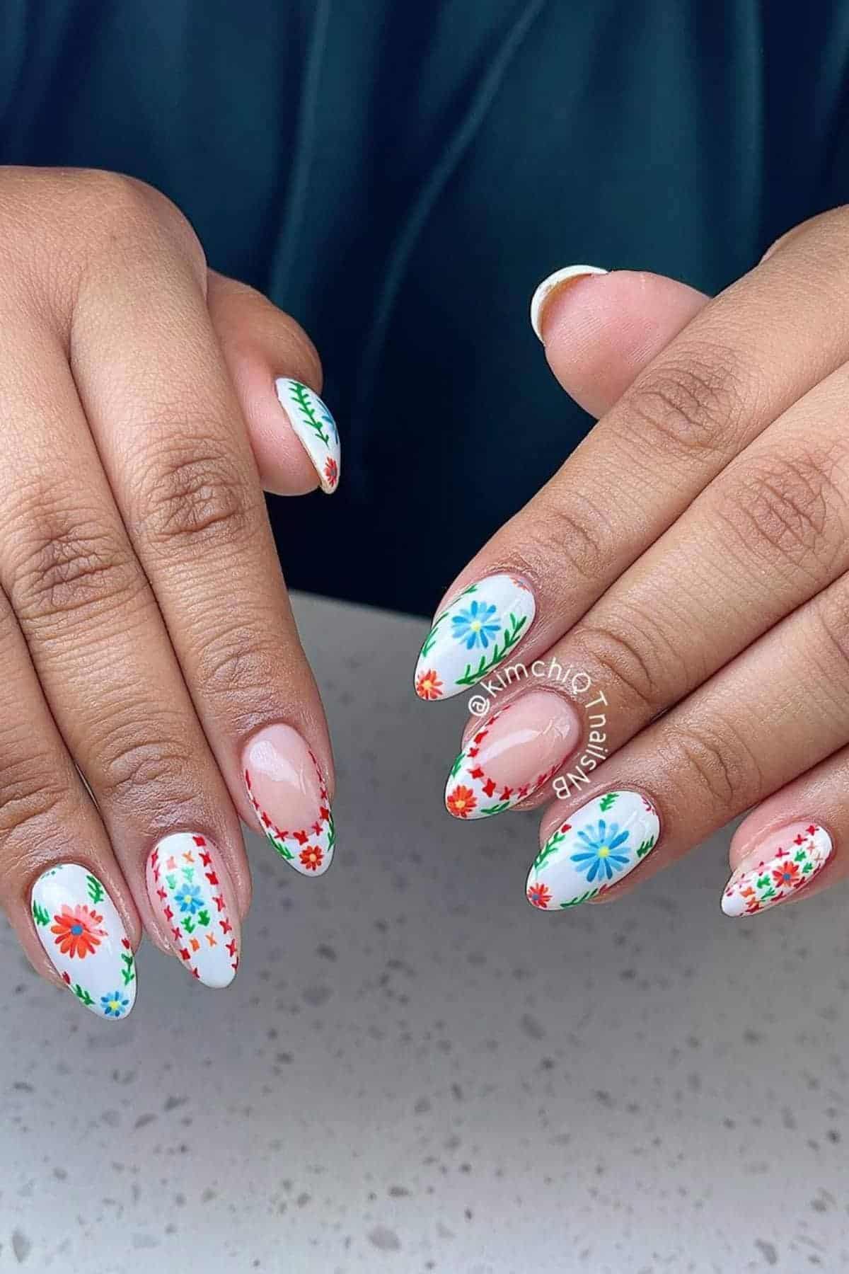 Hands with almond-shaped nails featuring a white base and colorful floral designs in red, blue, and green on each nail.
