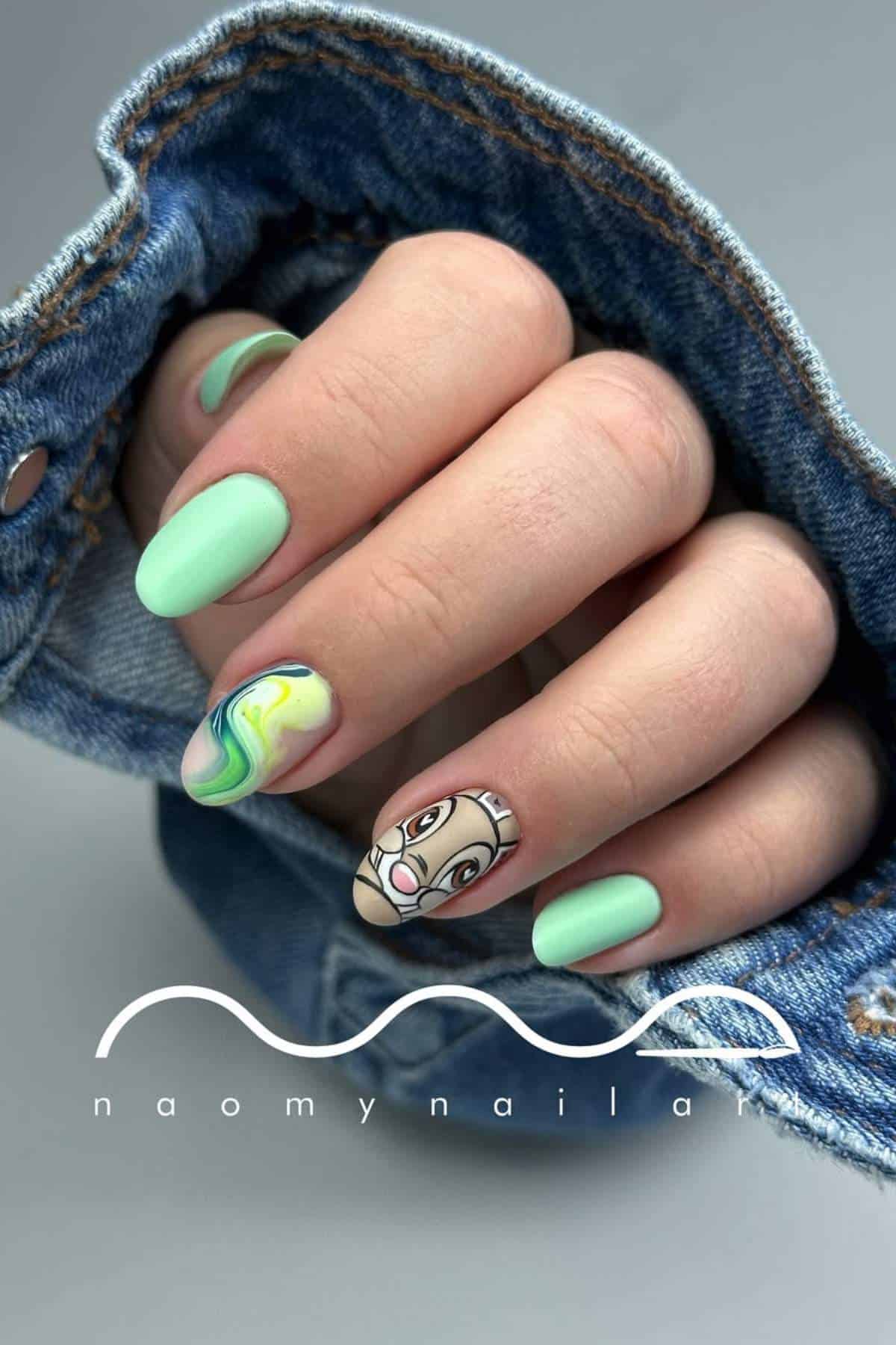 A hand with mint green nails, one nail with a marble swirl design, and one featuring a cartoon face, partially covered by a denim sleeve.
