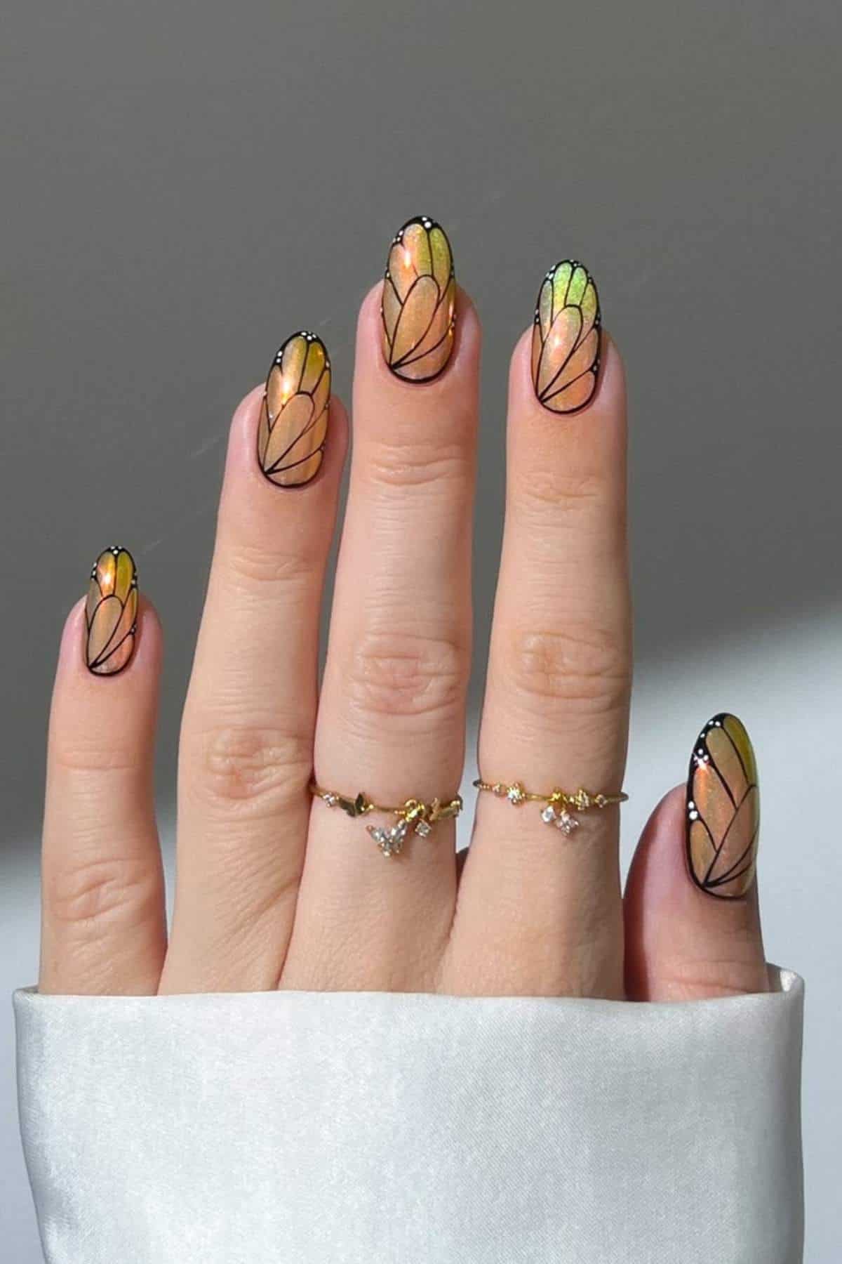 A hand with butterfly wing-inspired nail art in orange and yellow shades, outlined in black, wearing two delicate gold rings.
