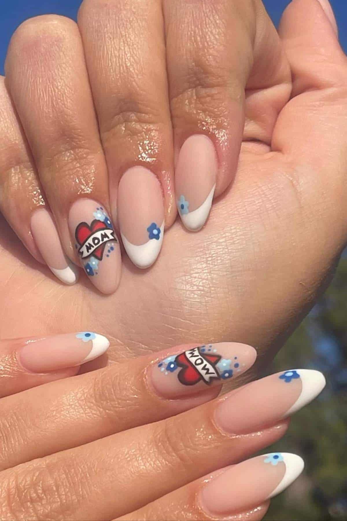 Hands with almond-shaped nails featuring nude polish, white French tips, blue flower designs, and heart tattoos with "MOM" text on two accent nails.