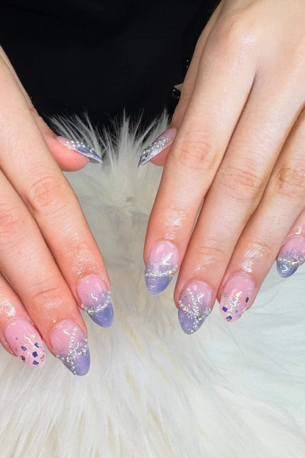 Hands with almond-shaped nails featuring a pastel purple French tip design, glitter accents, and iridescent flakes, displayed on a white furry surface.