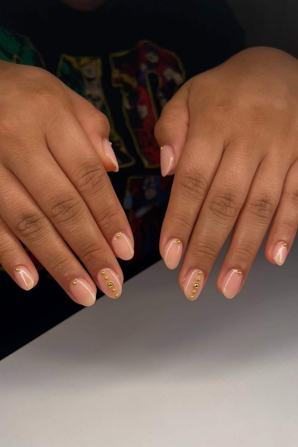 Two hands with light pink, almond-shaped nails featuring small gold studs near the cuticles on select fingers, resting on a white surface.
