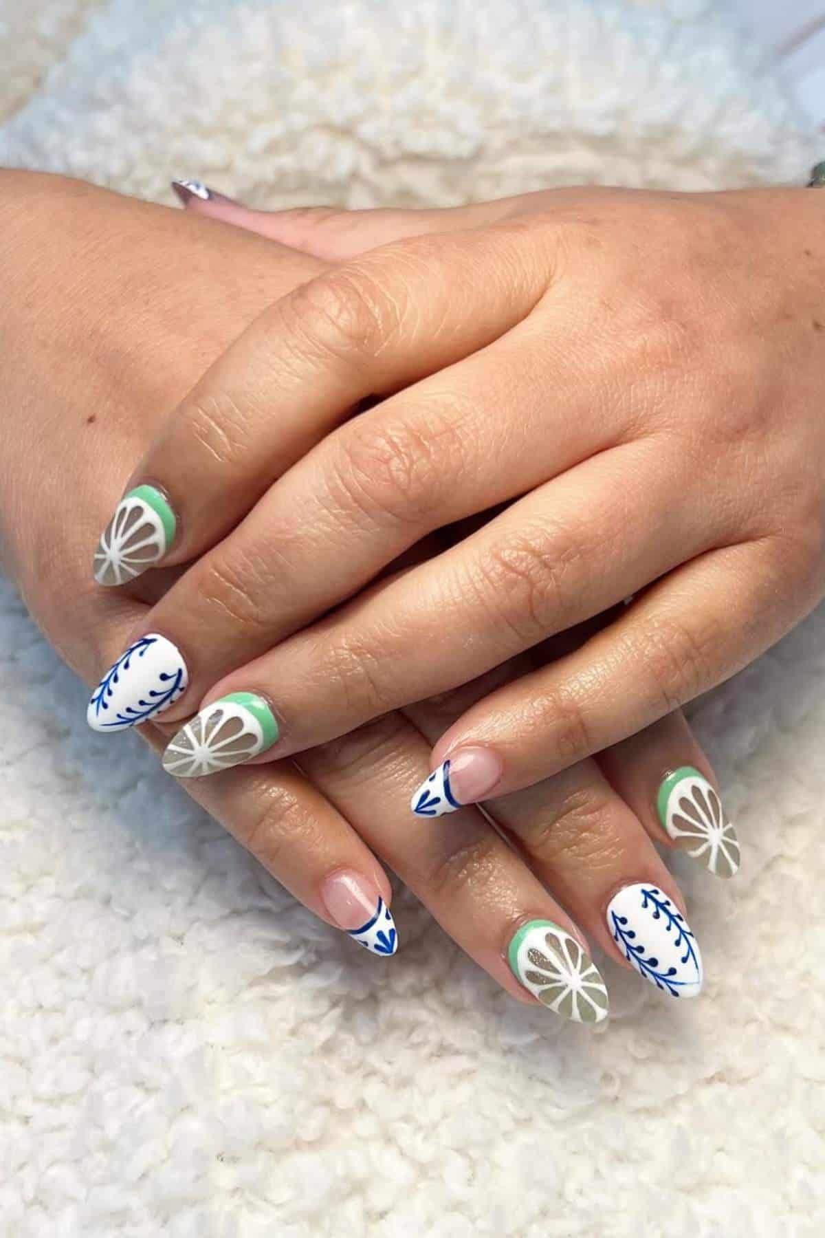 Hands with almond-shaped nails featuring a white base and blue, green, and beige botanical and geometric designs.