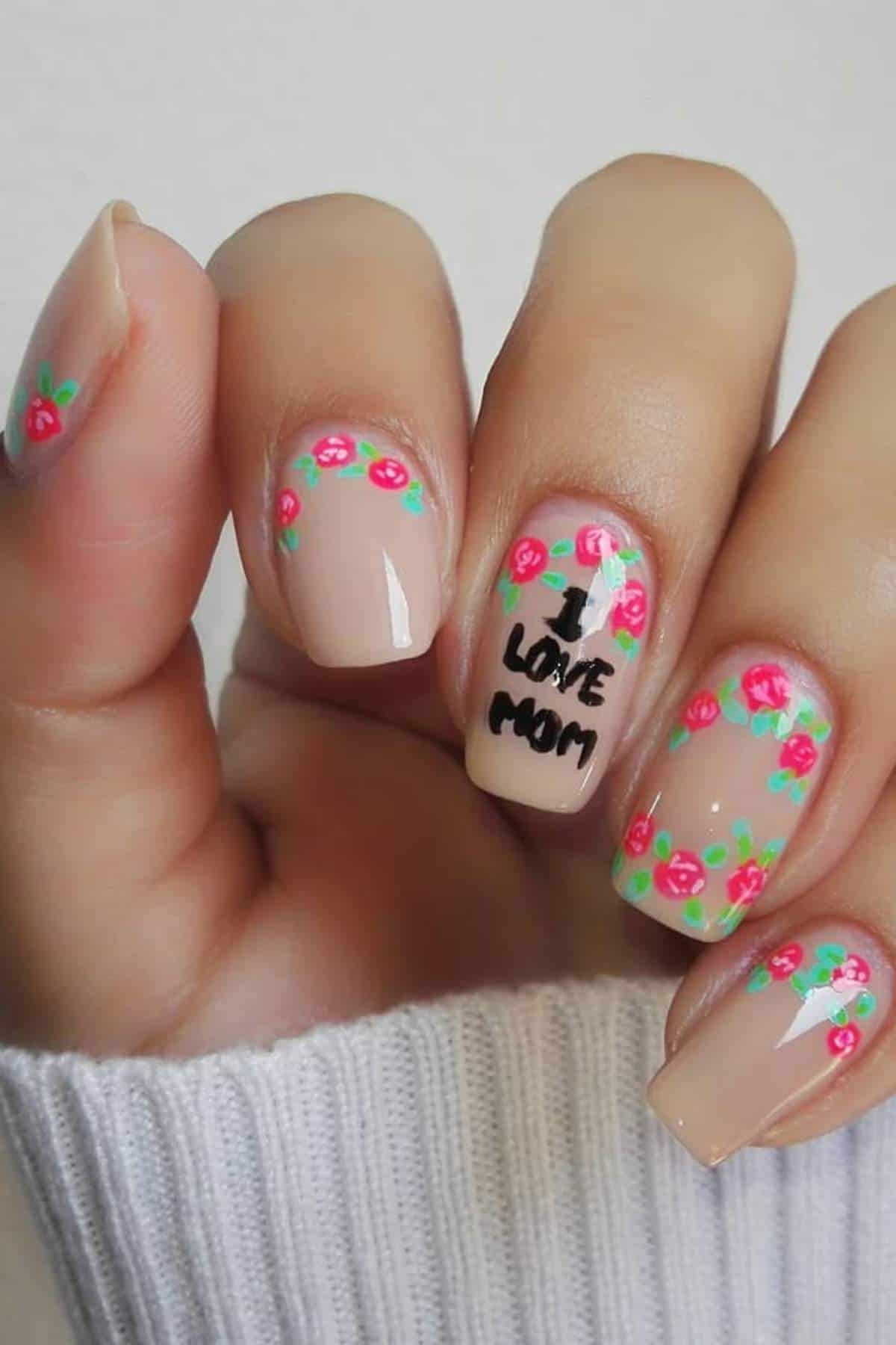 A hand with light nude nail polish, small pink flower designs on each nail, and "I love mom" written in black on the ring fingernail.
