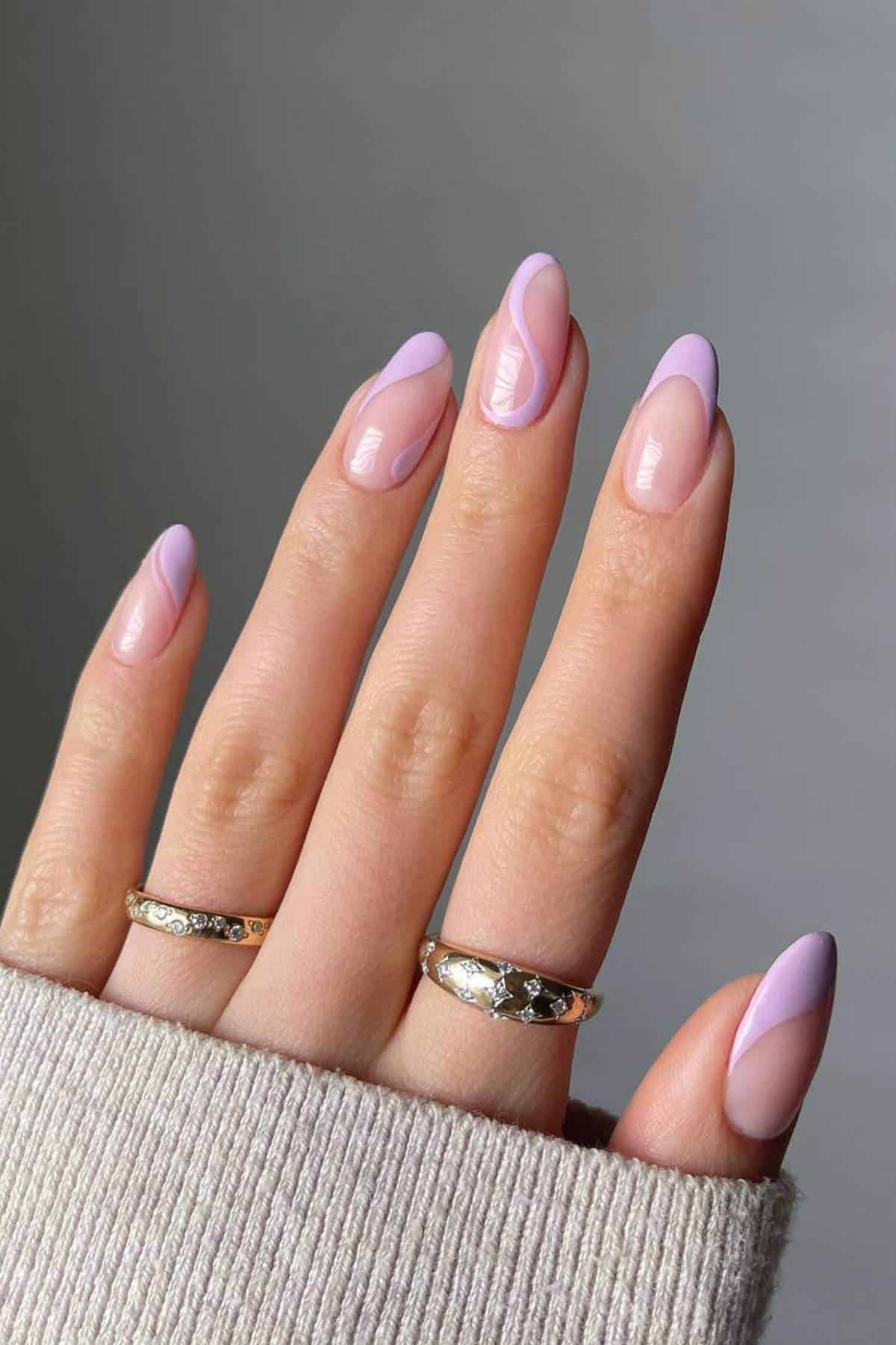 A hand with almond-shaped nails painted in a pastel pink and lavender swirl design, wearing two gold rings, partially covered by a beige sweater.