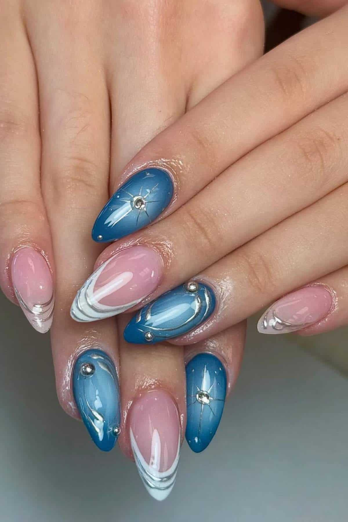Hands with manicured nails featuring a mix of blue and pink designs, white accents, and small silver studs on almond-shaped nails.