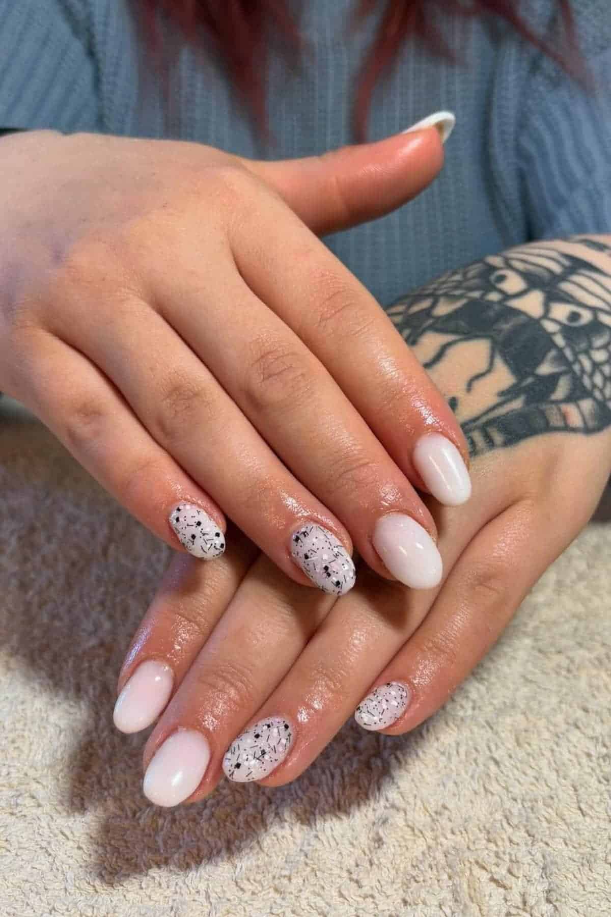 A person with light pink oval-shaped nails, some featuring black speckled designs, rests their hands on a towel; a tattoo is visible on one wrist.