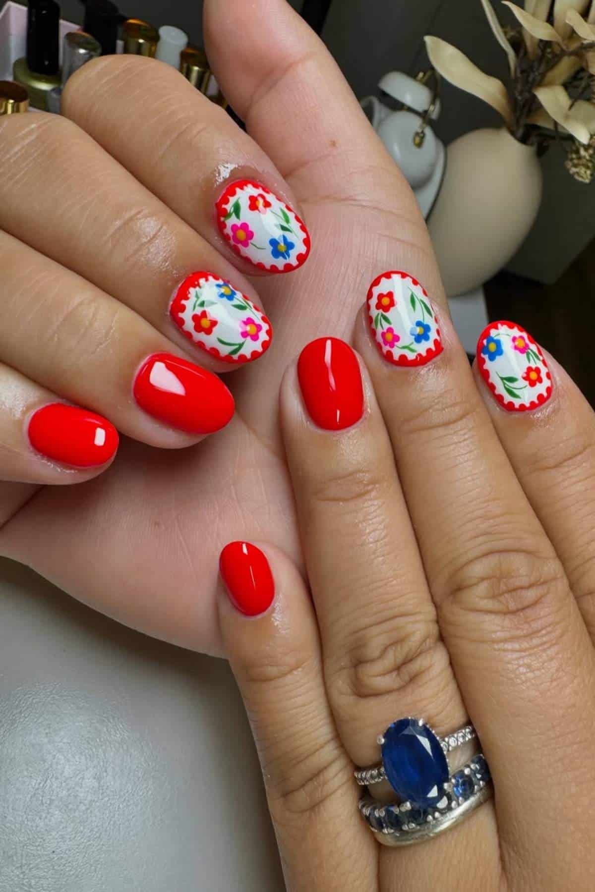 Hands with red nail polish; some nails feature a white base with colorful floral designs and red scalloped edges. A ring with a blue stone is worn on one finger.