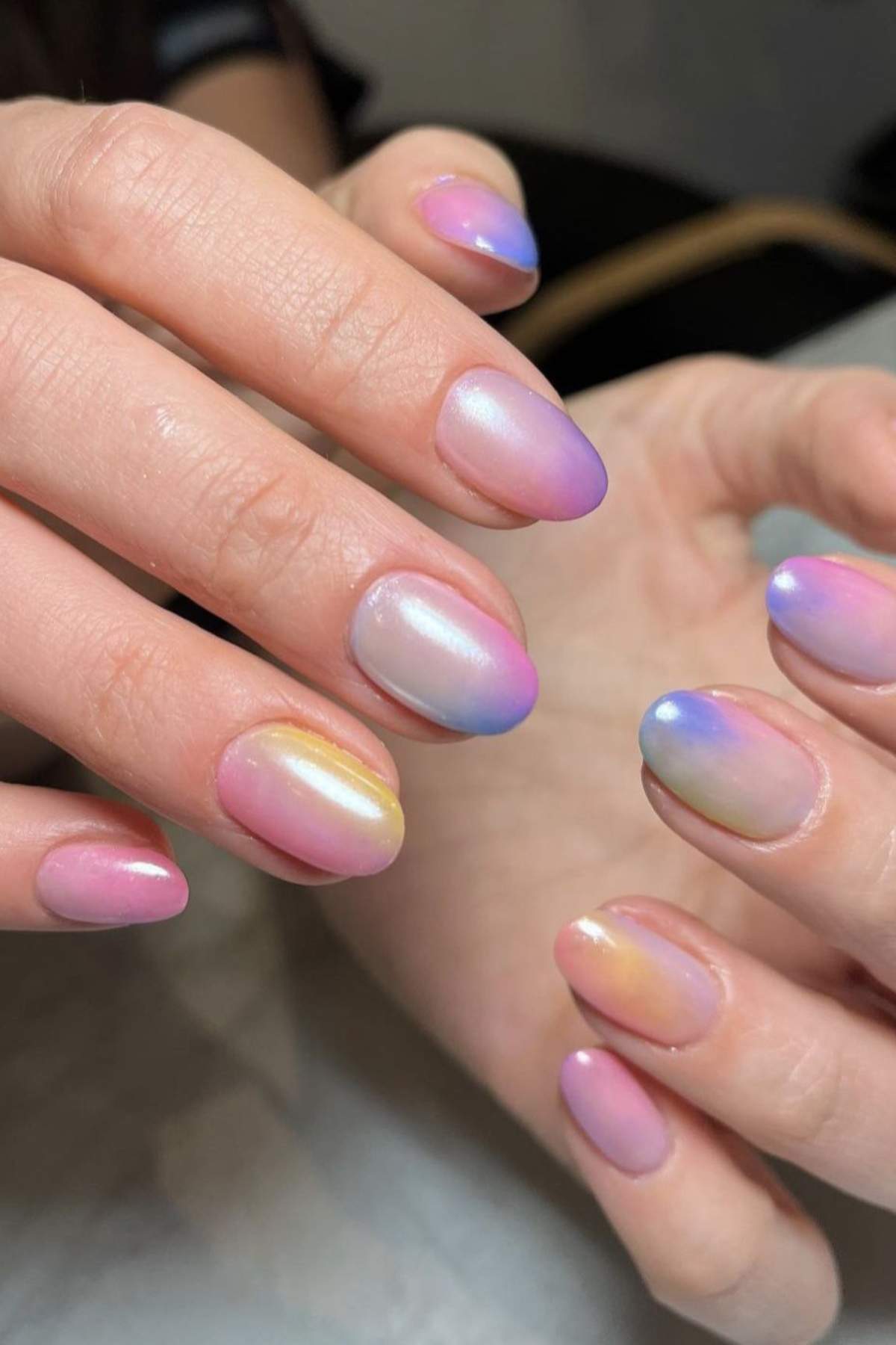 Close-up of hands with short, oval-shaped nails featuring a pastel, iridescent gradient nail polish in shades of pink, blue, yellow, and purple.