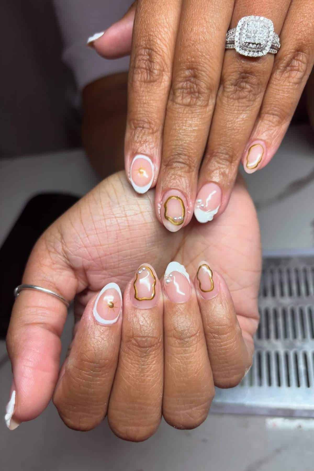 Close-up of hands with almond-shaped nails featuring gold-lined abstract designs and white tips; person wears silver rings, including a diamond-studded ring.