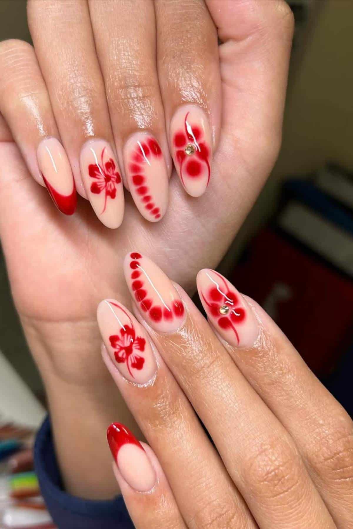 Hands with long almond-shaped nails featuring a nude base, red floral designs, red french tips, and small rhinestone accents on some nails.
