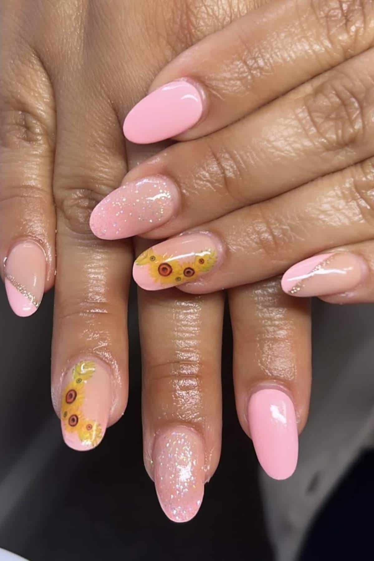Hands with almond-shaped nails painted light pink; some nails have glitter and sunflower designs on the tips.