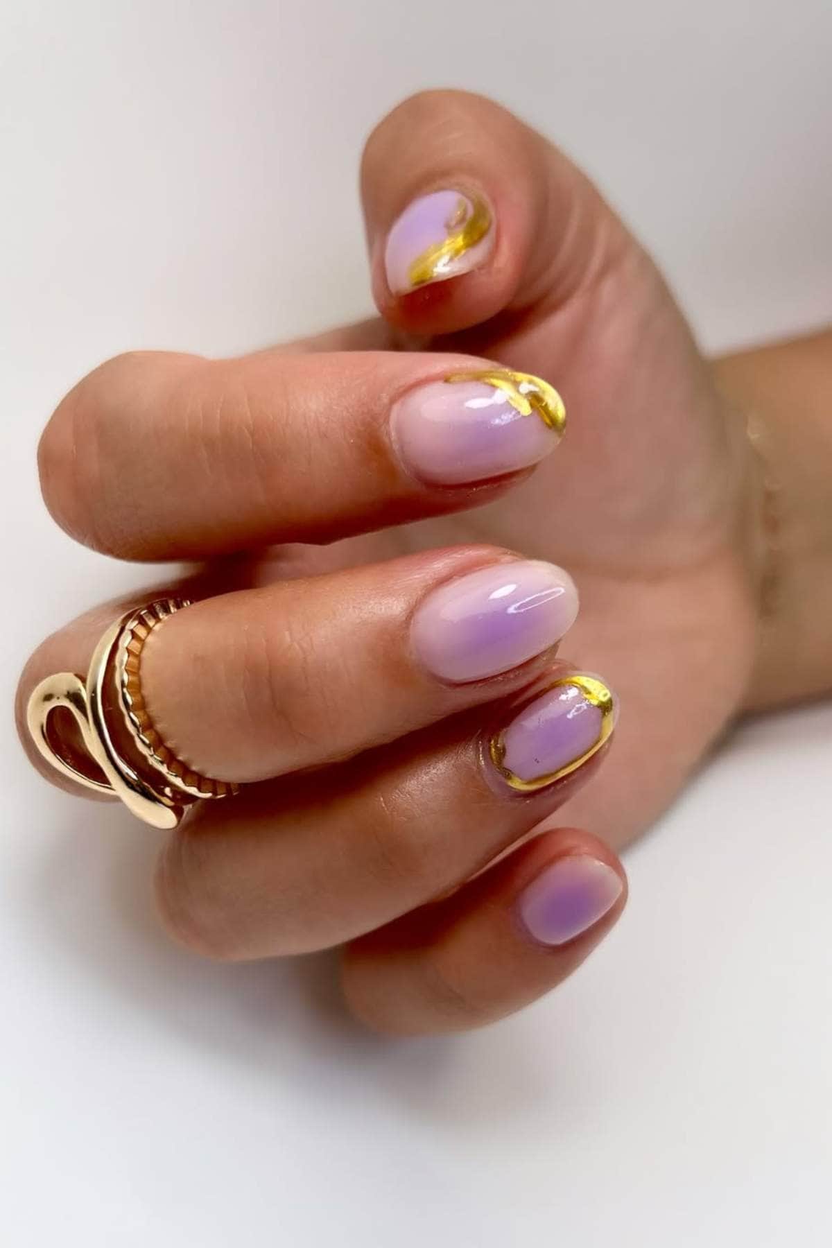 A hand with short, oval nails painted in a gradient of pink and purple, featuring gold accents near the cuticles. The person wears gold rings on their fingers.
