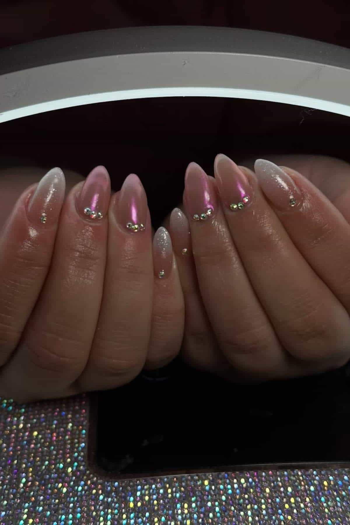 Hands with long, almond-shaped nails featuring a pink and white ombre design, decorated with small gold bead accents, shown under a bright lamp.